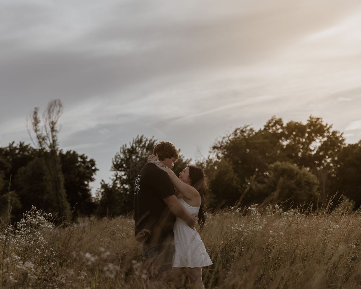 A couple embraces and kisses in a golden wildflower field at sunset during an outdoor engagement session.