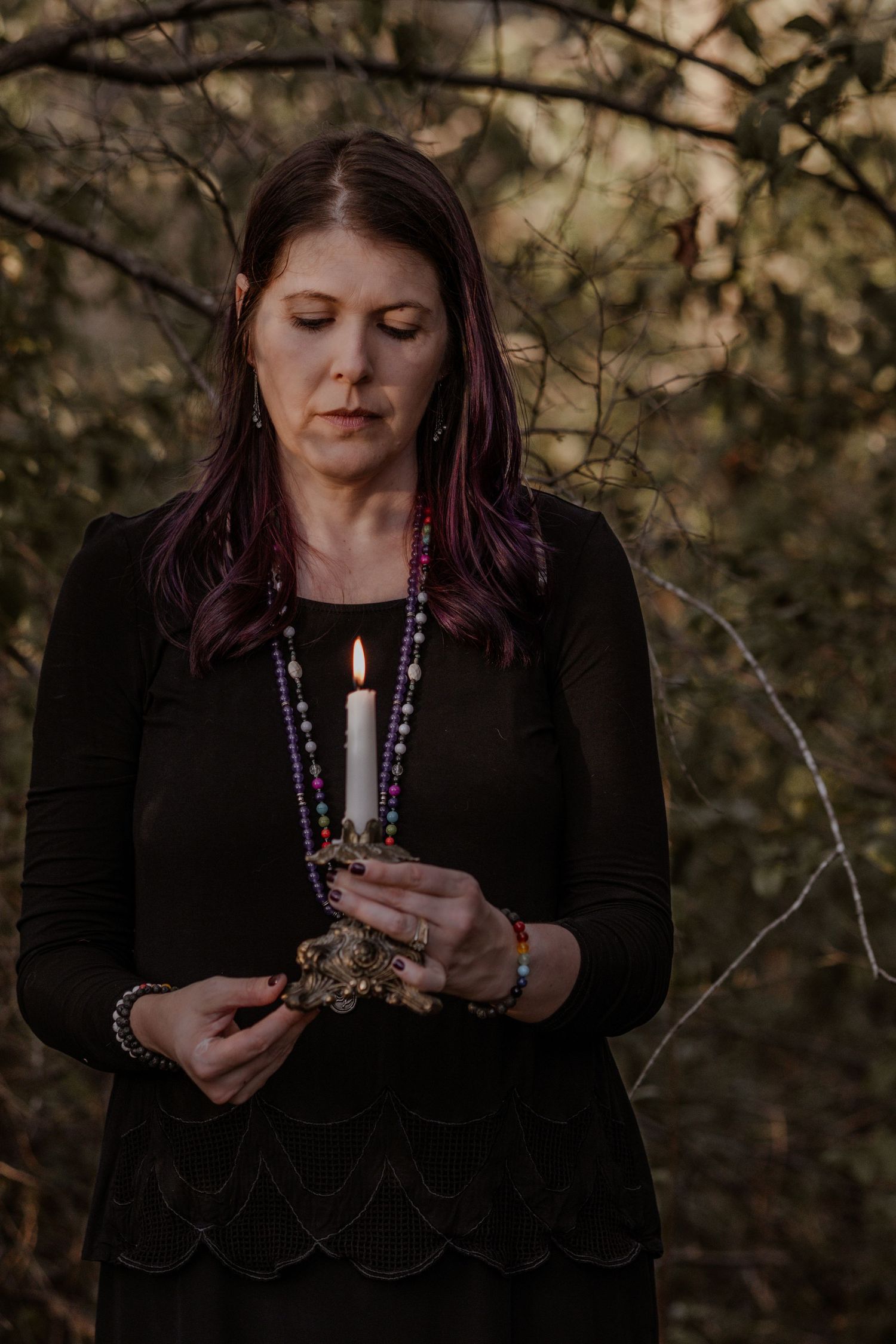 Person in black holding candles and wearing purple beaded necklace in outdoor setting.