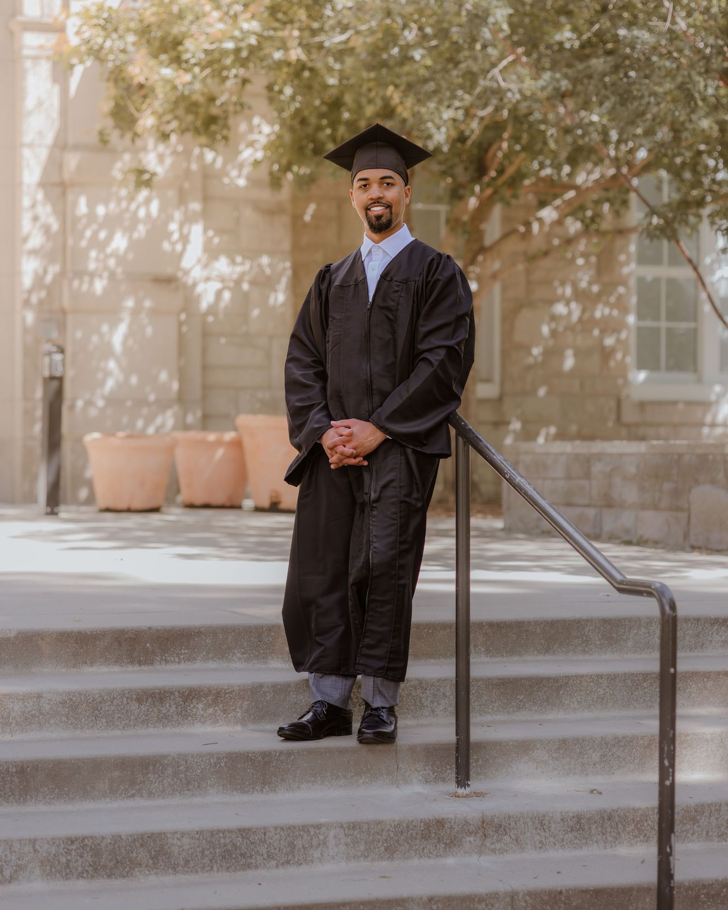 Graduate in black cap and gown stands on steps outdoors, smiling confidently at camera.