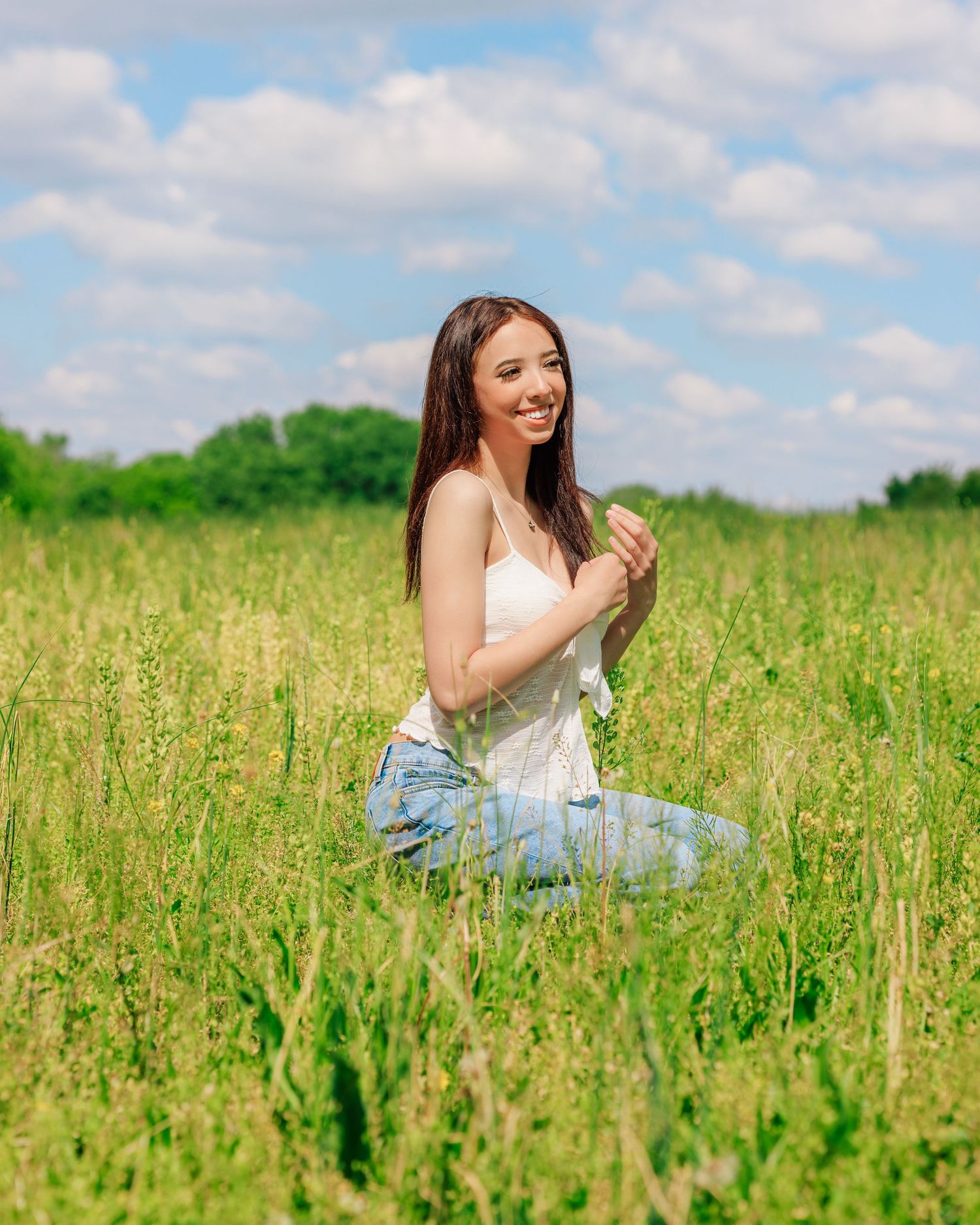 Young woman sitting in a lush green meadow, smiling joyfully, wearing a white top and jeans on a sunny day.