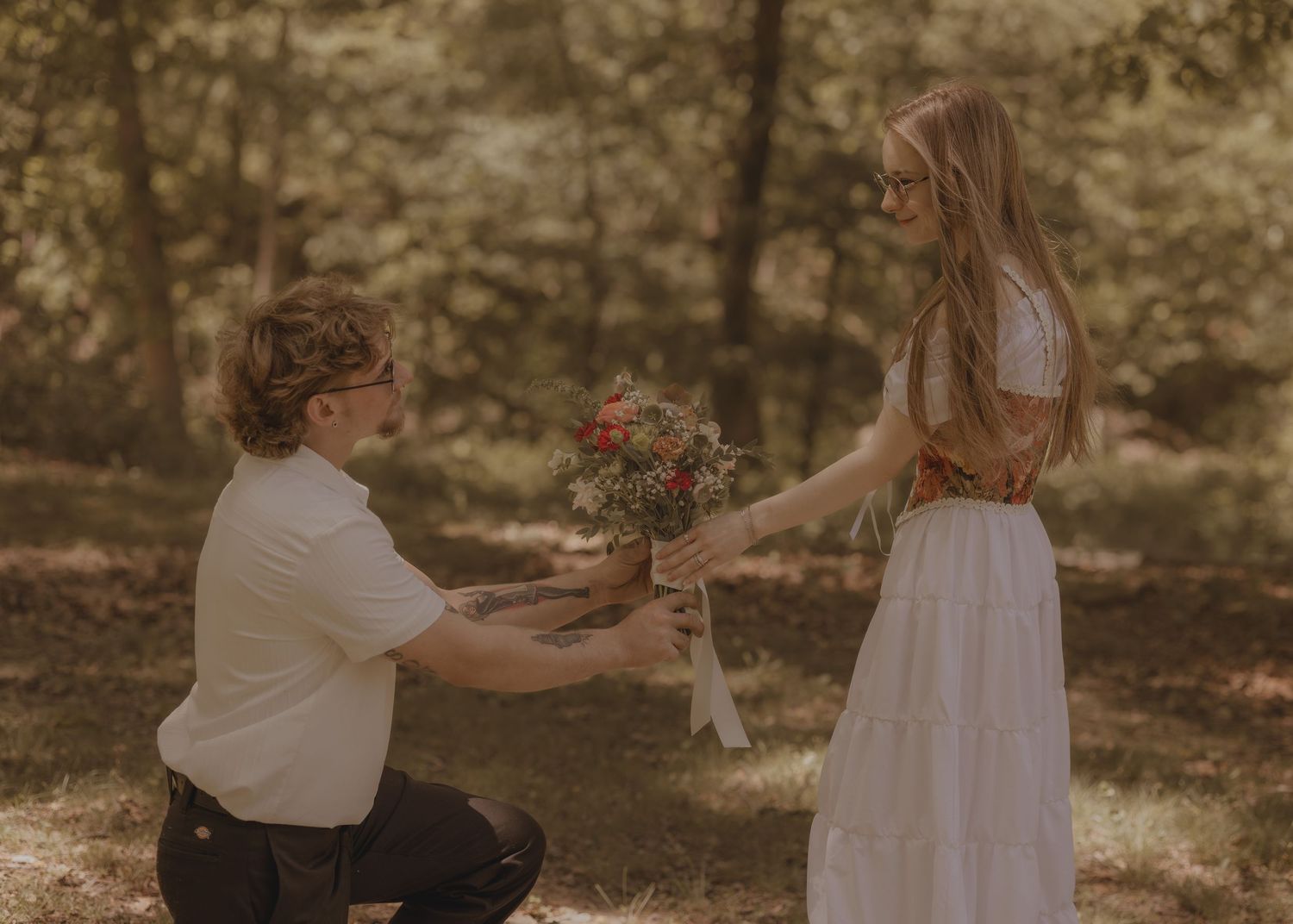Man kneeling on one knee presenting a bouquet of flowers to a woman in a white dress outdoors.