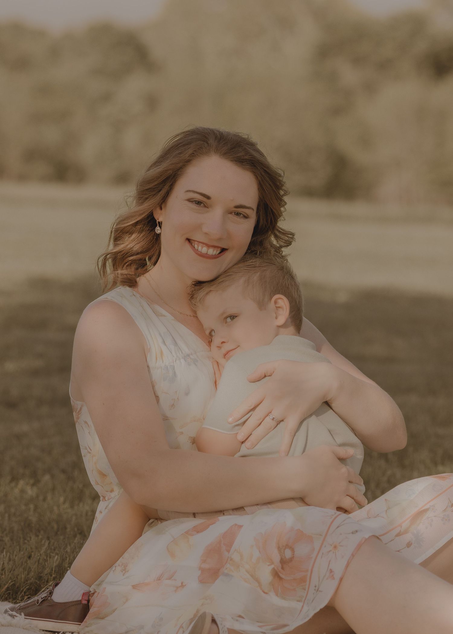 Smiling mother holding sleeping baby outdoors on grass in floral dress during golden hour photography session.