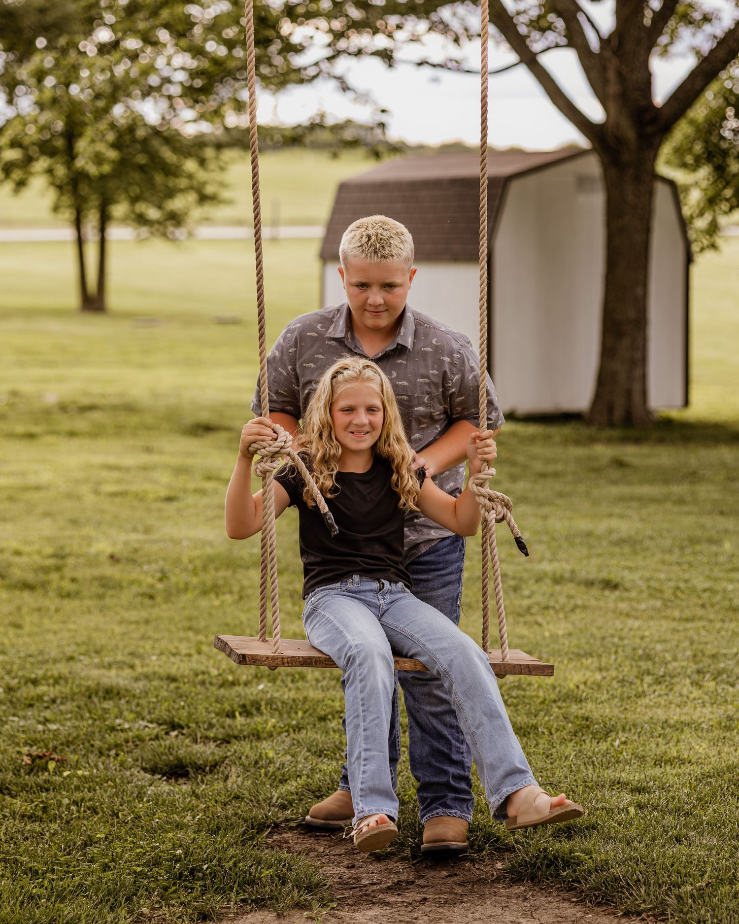 Two kids playing on a rope swing in a green backyard near a shed.