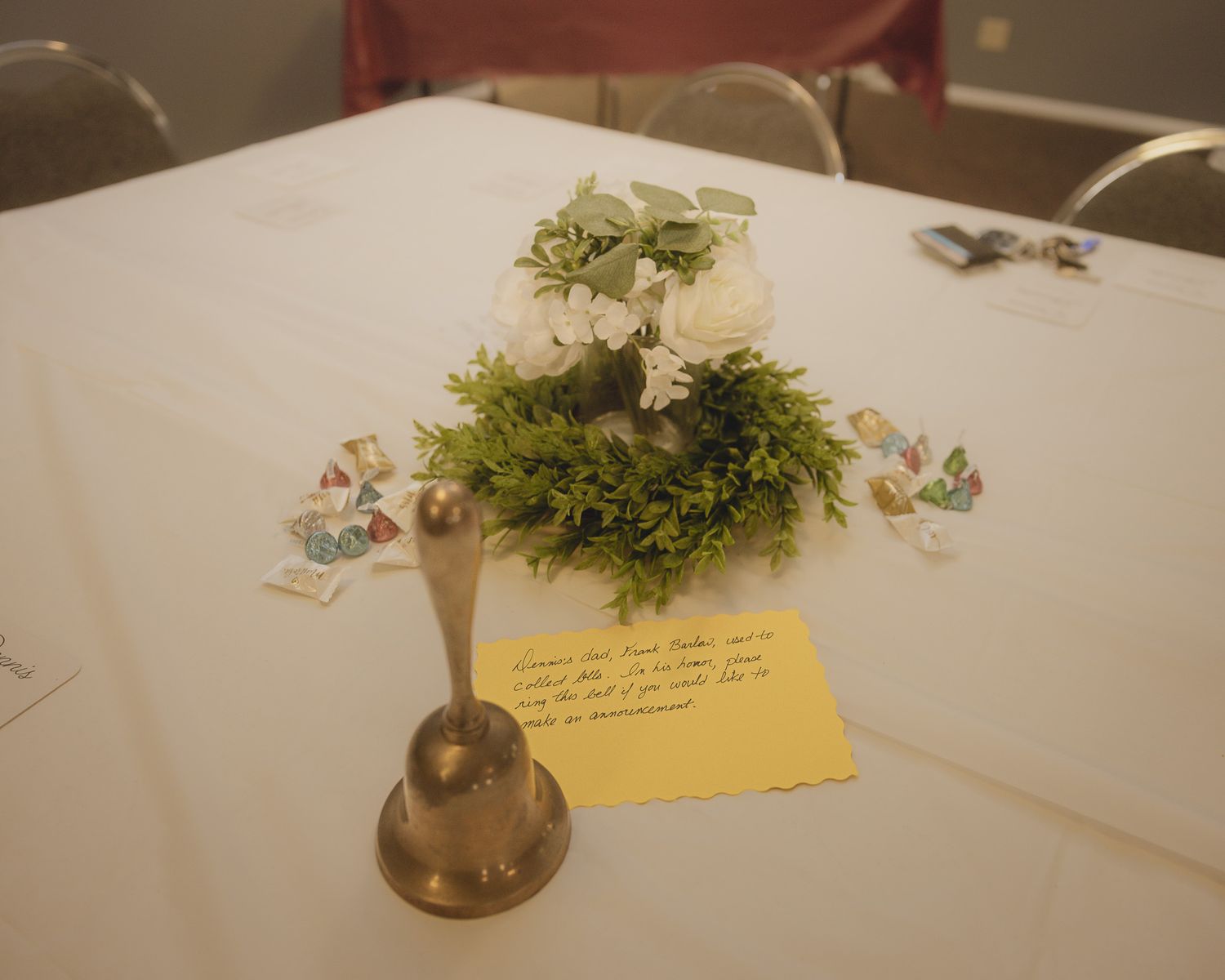 A decorative centerpiece with greenery and flowers sits atop a white tablecloth with a small note card.