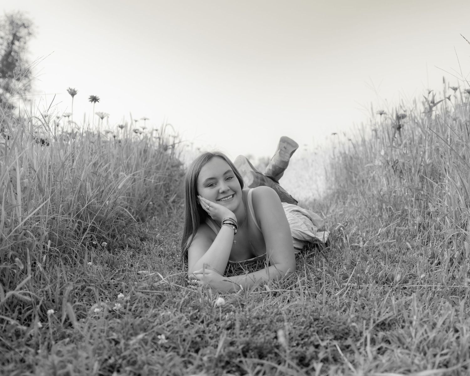 Black and white photo of a smiling young woman lying in a grassy meadow with wildflowers.