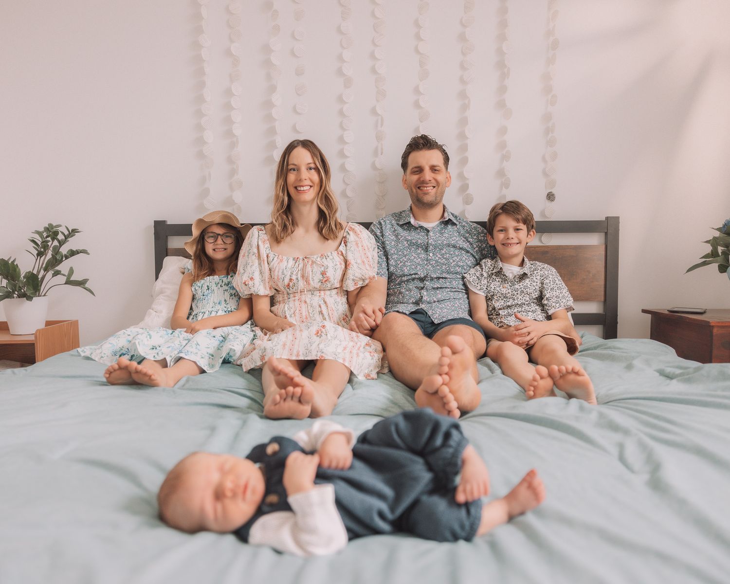 Smiling family of five poses on a bed with newborn baby in the foreground.
