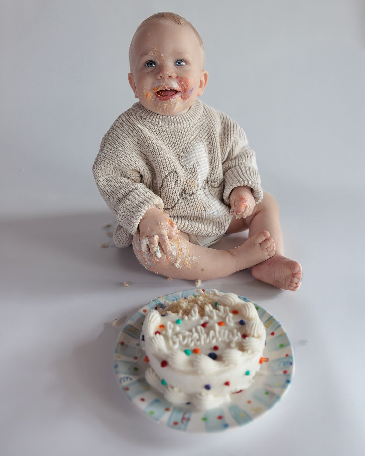 Happy baby with frosting on face smashes first birthday cake while wearing a beige knit sweater romper.