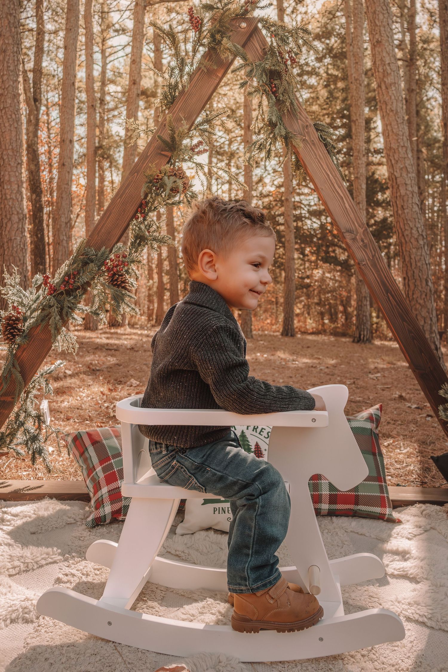 A small child sits on a white wooden bench in a sunlit forest setting with pine trees in the background.