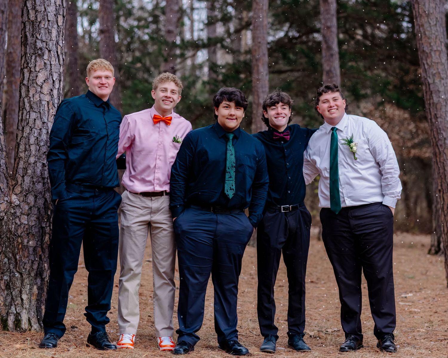 A group of groomsmen in navy suits and dress shirts pose together in a forest setting during a wedding celebration.