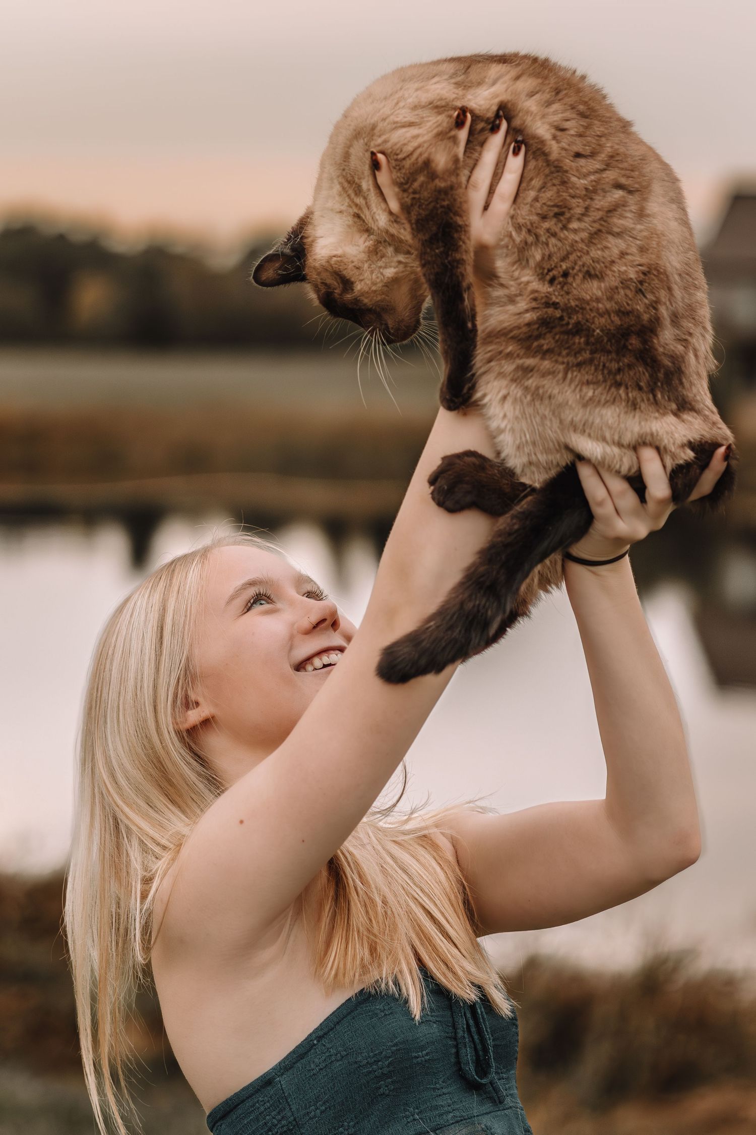A person lifts and plays with their brown cat against a sunset backdrop near a lake.