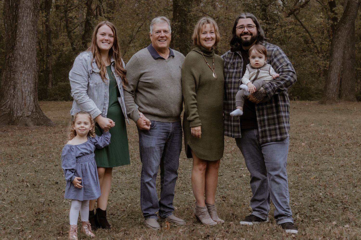 A group of people stand together outdoors in casual clothing for a family portrait in a wooded setting.