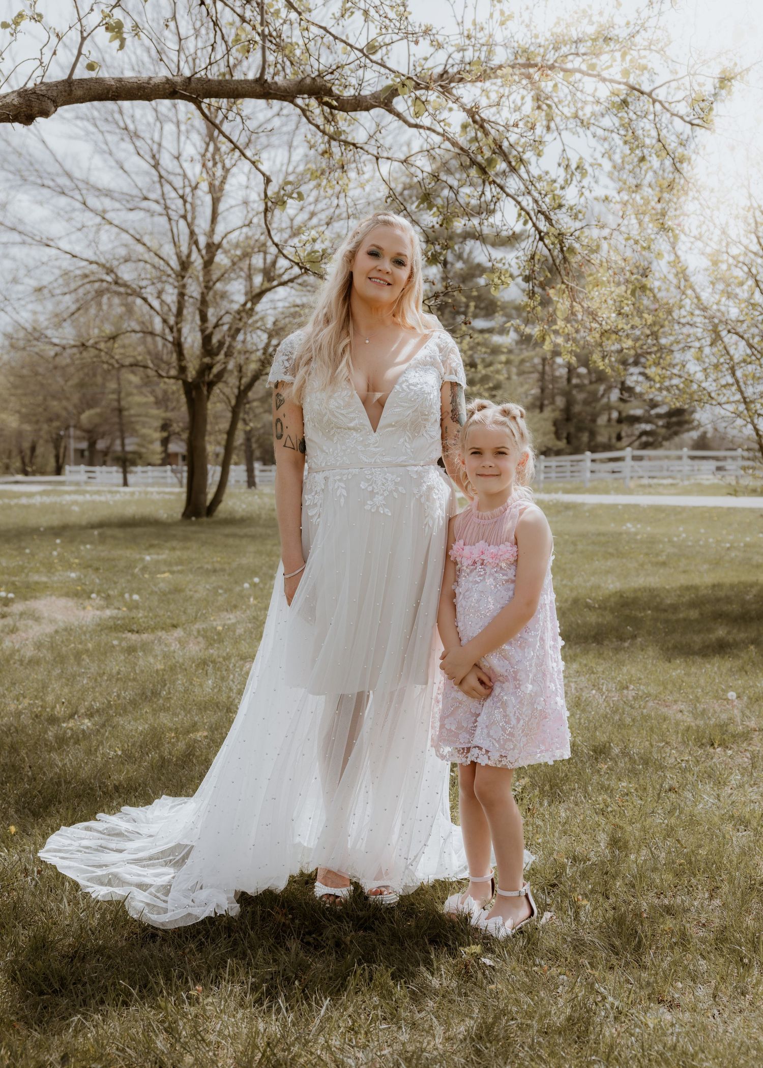 Bride in lace wedding dress posing with young flower girl in pink dress outdoors.