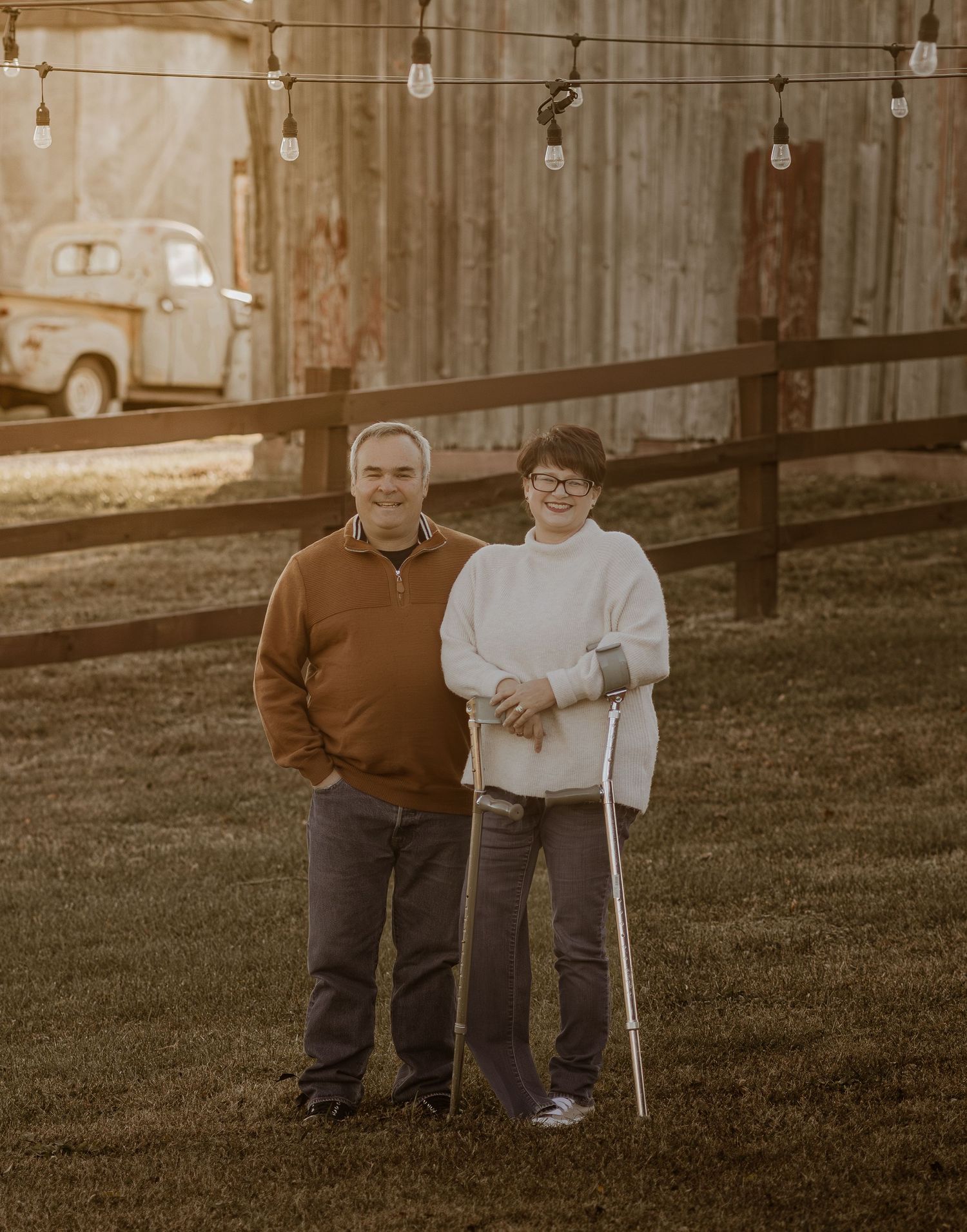 Couple smiling outdoors near rustic wooden fence with string lights, woman using a crutch, vintage truck in background.