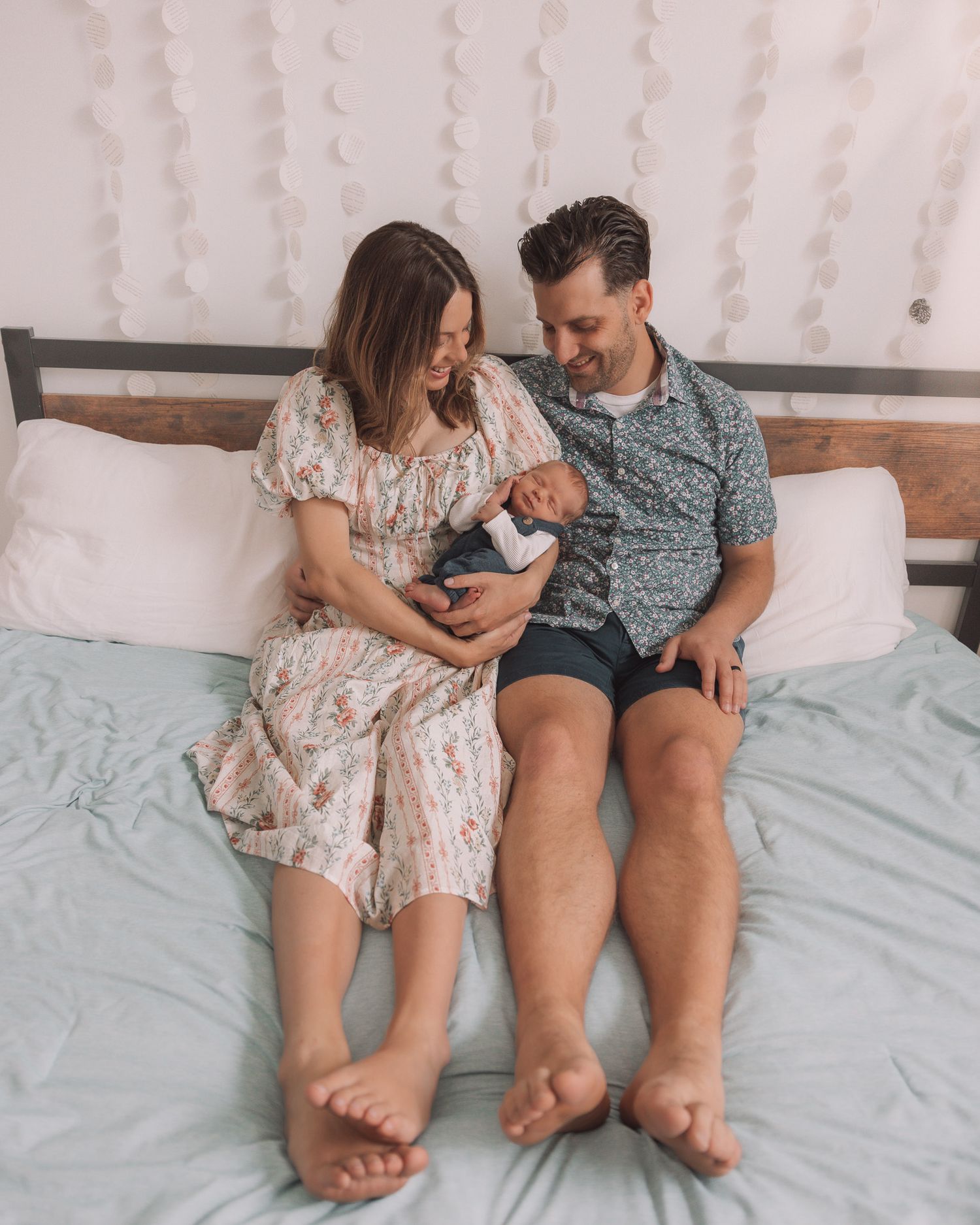 A couple sitting on a bed looking down at their newborn baby, woman in floral dress, man in blue shirt.