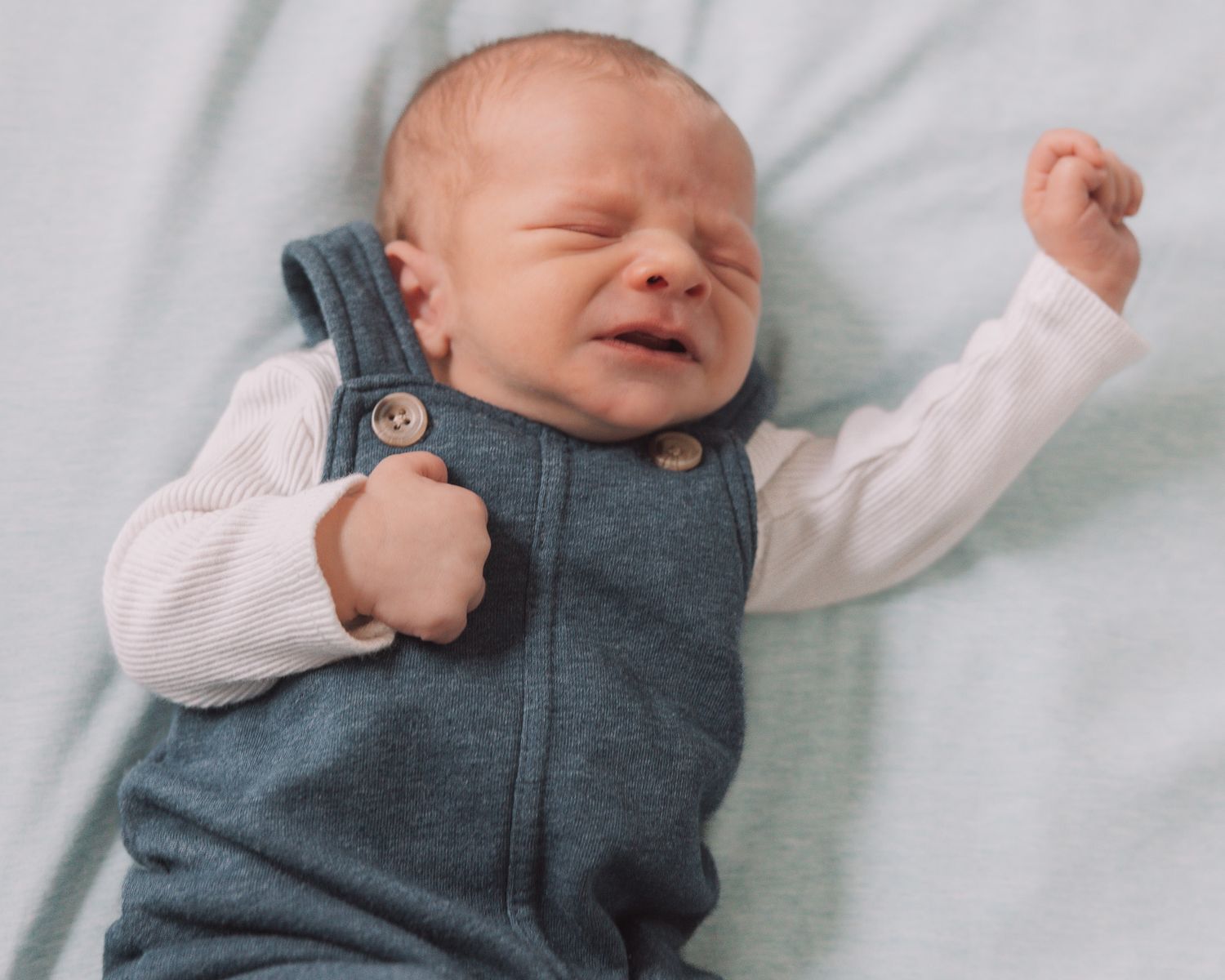 Newborn baby in a teal romper lying on a light blue blanket with tiny fists raised.