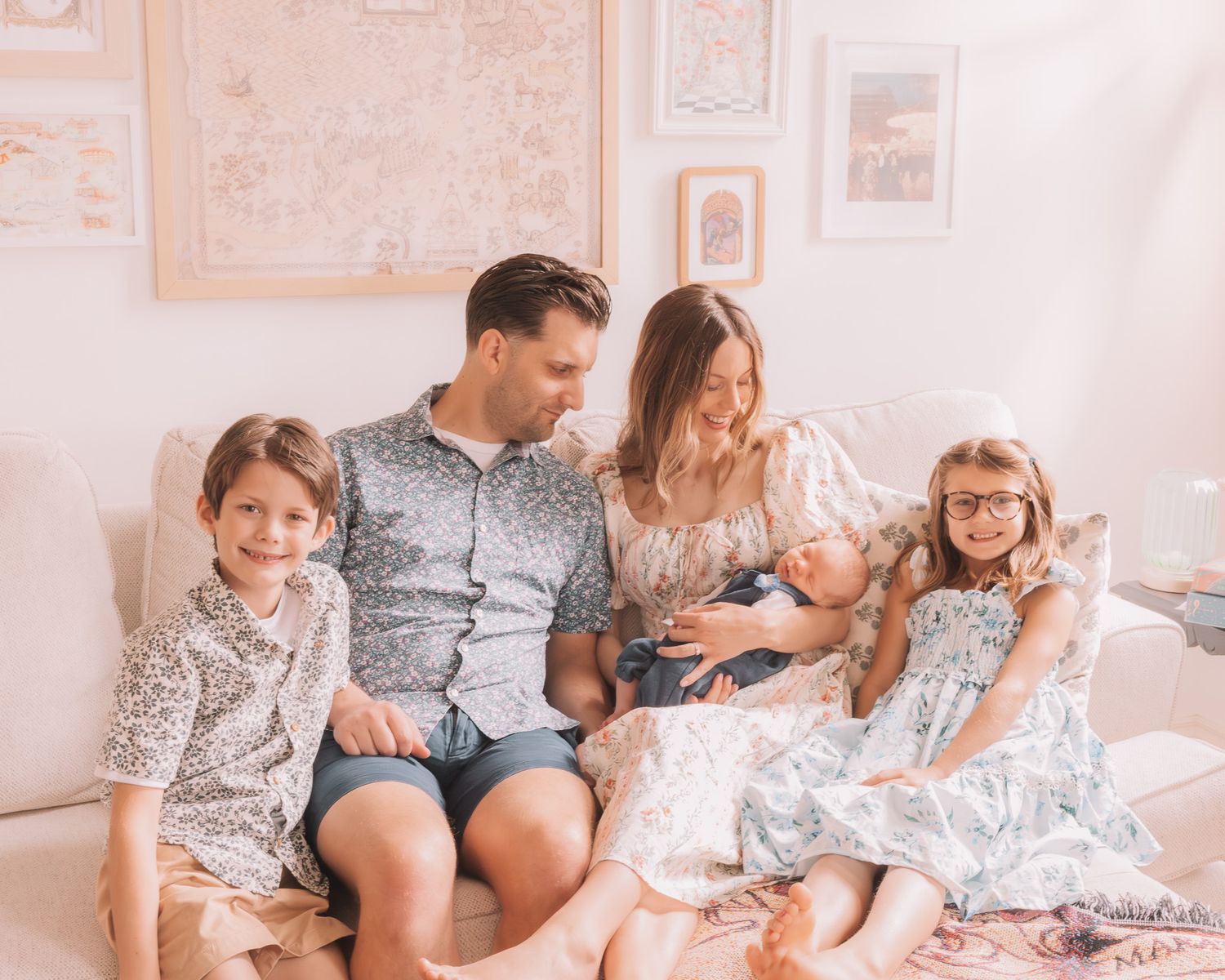 Happy family of five sitting on a couch, parents with three young children including a newborn, in a bright cozy room.