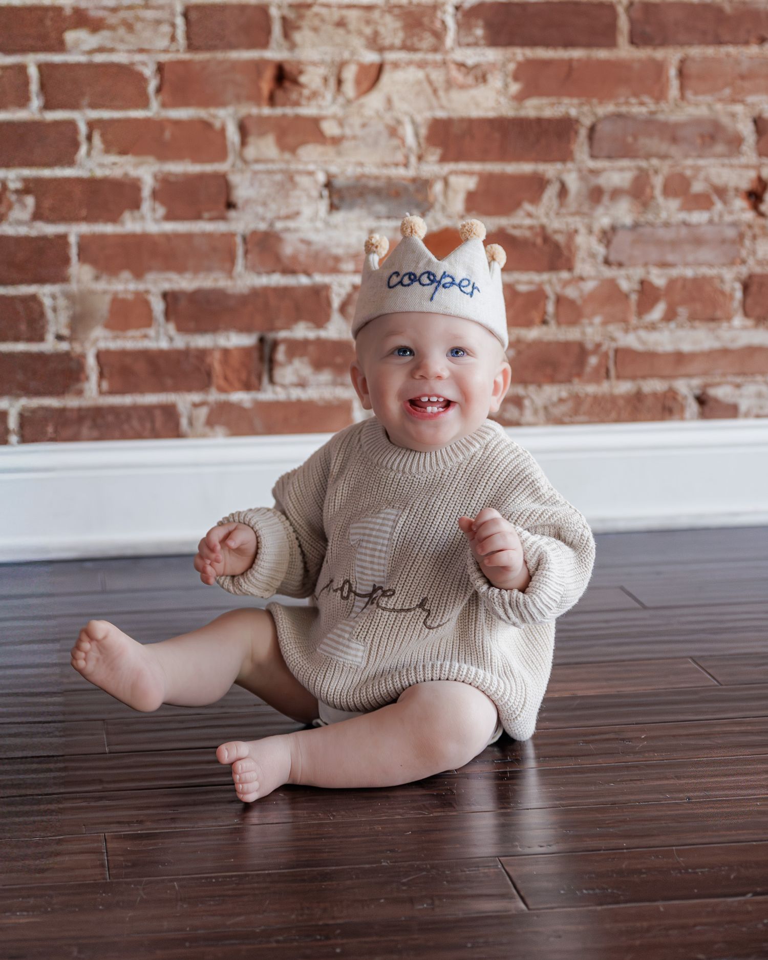 Baby named Cooper sits on hardwood floor wearing knit romper and personalized crown hat, smiling at camera.