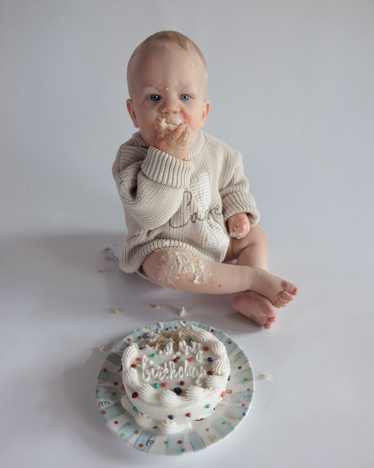 Cute baby in knit sweater eating birthday smash cake with frosting on face, colorful sprinkled cake in front.