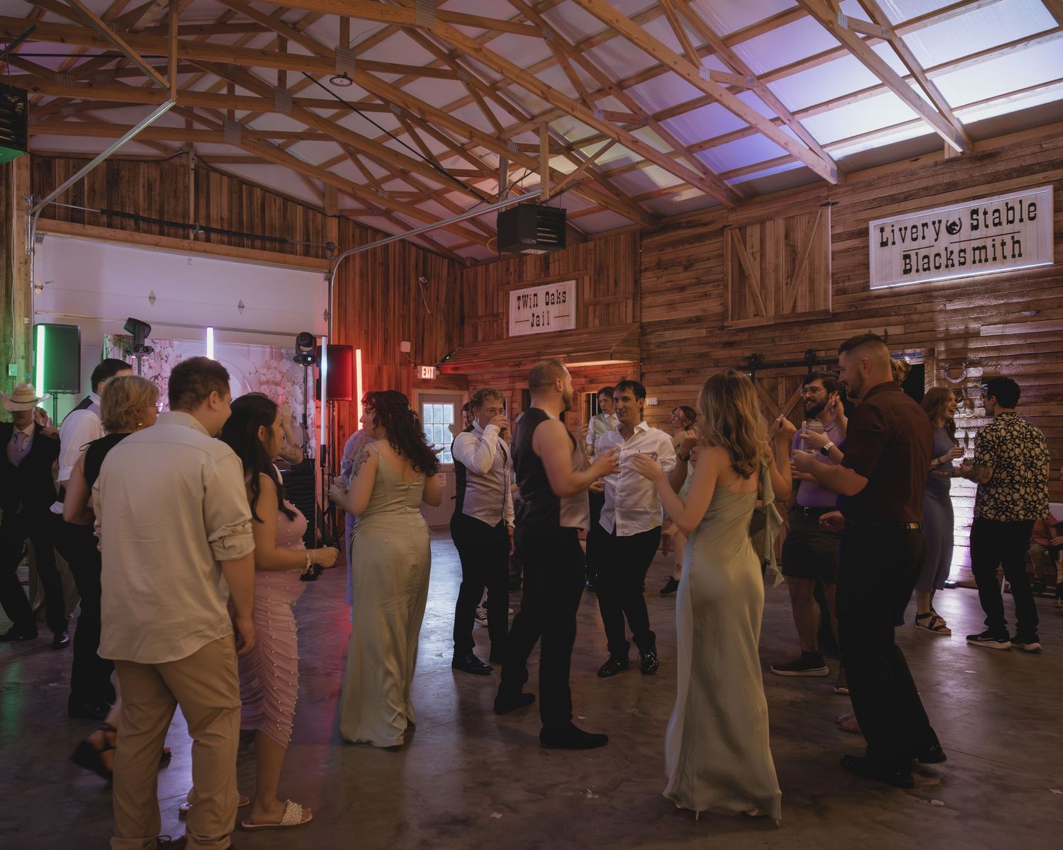 Wedding reception guests dancing and socializing inside a rustic barn venue with warm atmospheric lighting.