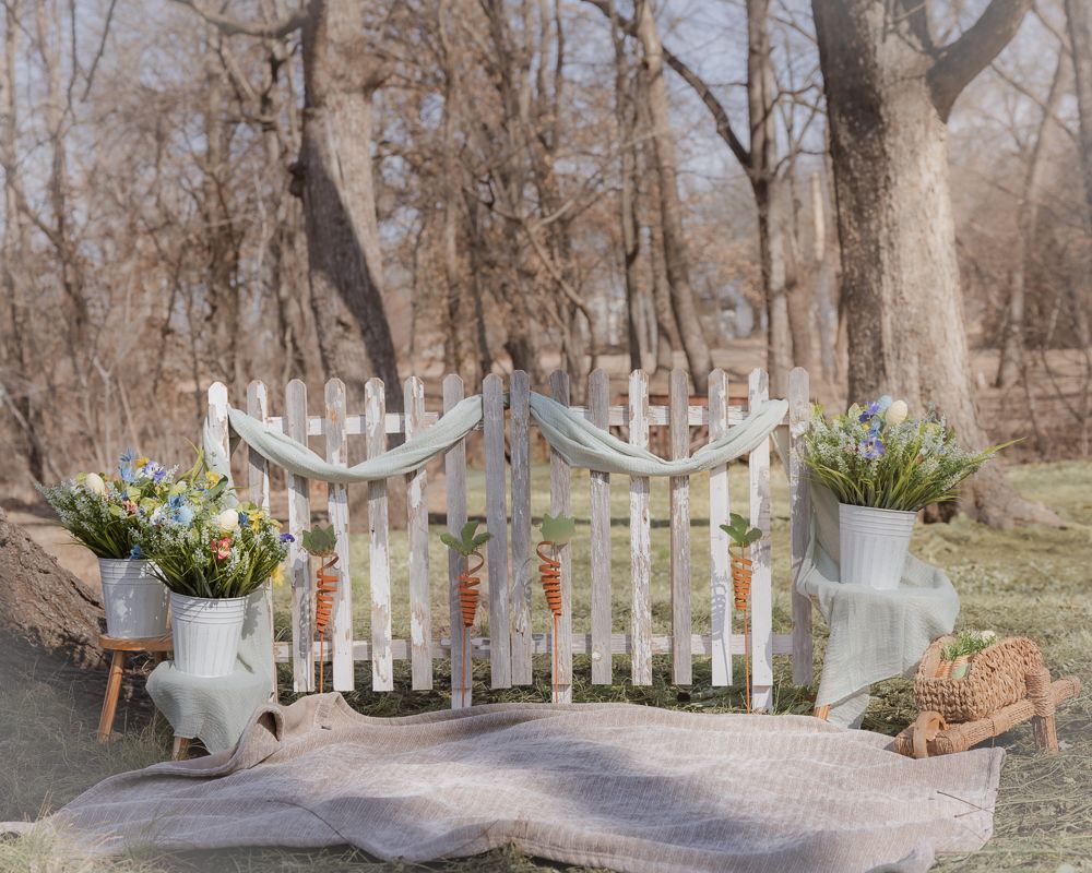 A white picket fence gate leads to a winding path through bare winter trees with a rustic garden setting.