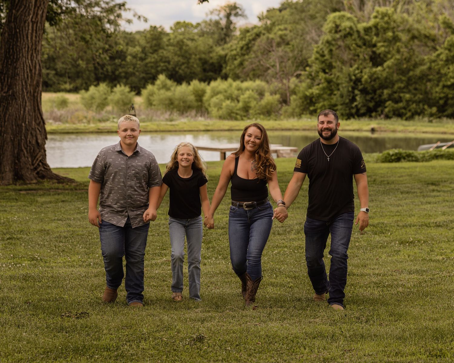 Happy family of four holding hands and walking together across a lush green lawn near a serene pond.