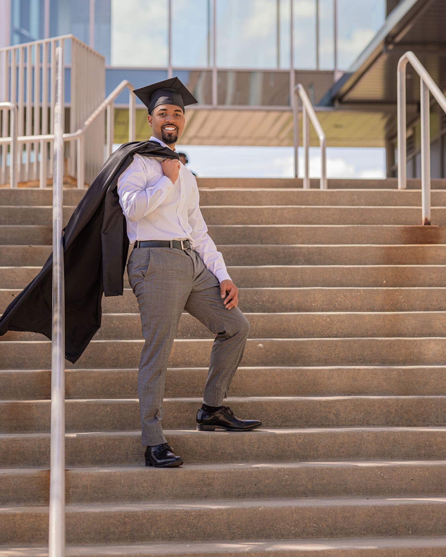 Smiling graduate poses on outdoor stairs holding black cap and gown over shoulder in formal attire.
