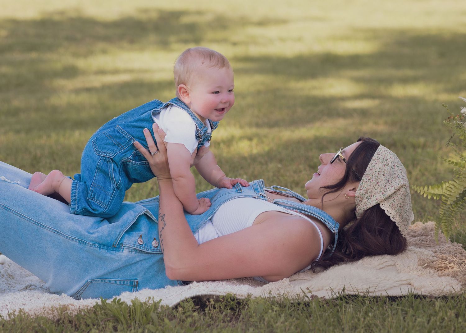 A mother lying on a blanket in the grass playfully lifting her smiling baby wearing denim overalls outdoors.