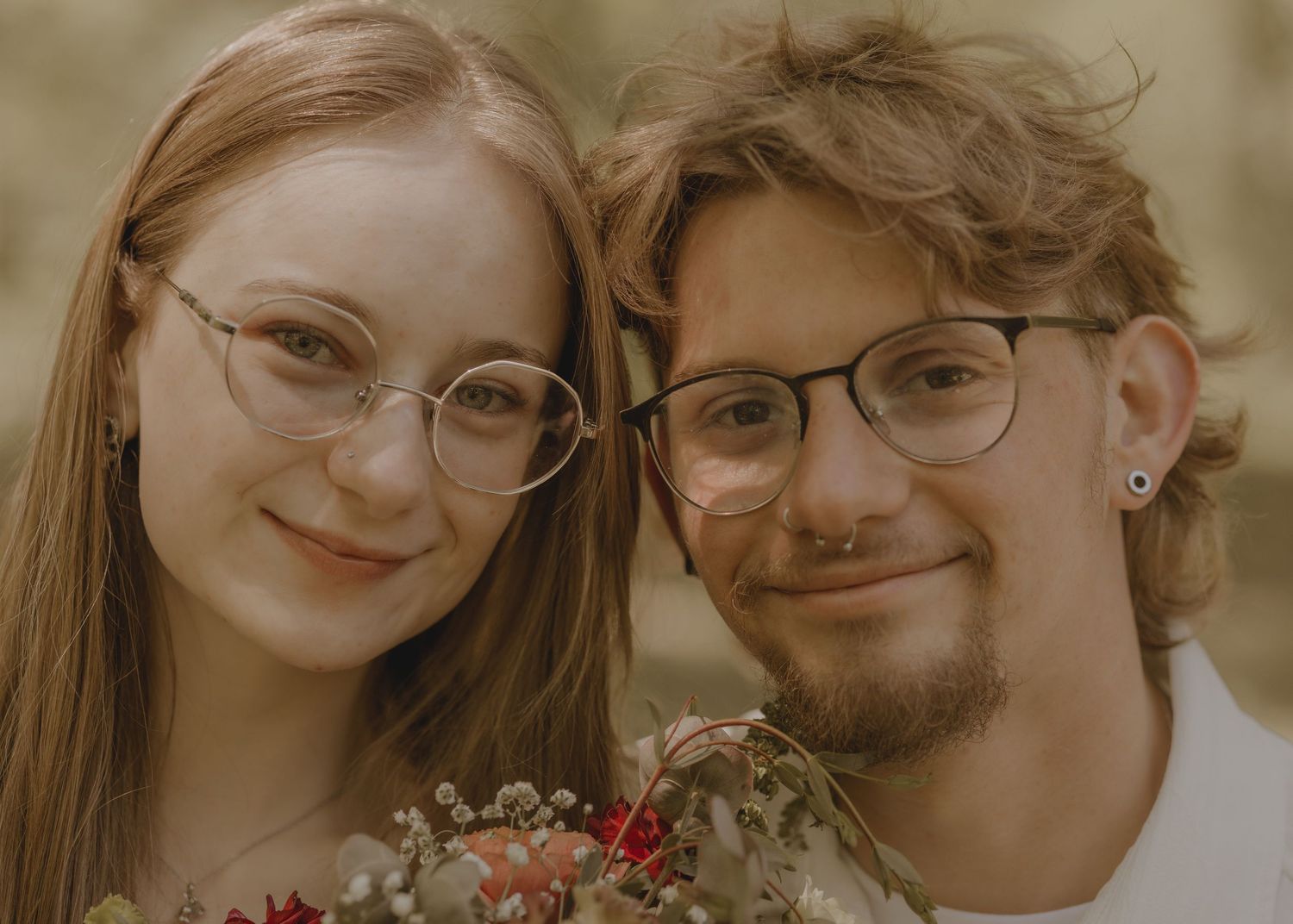 Young couple with glasses smiling closely together, holding a bouquet of flowers outdoors.