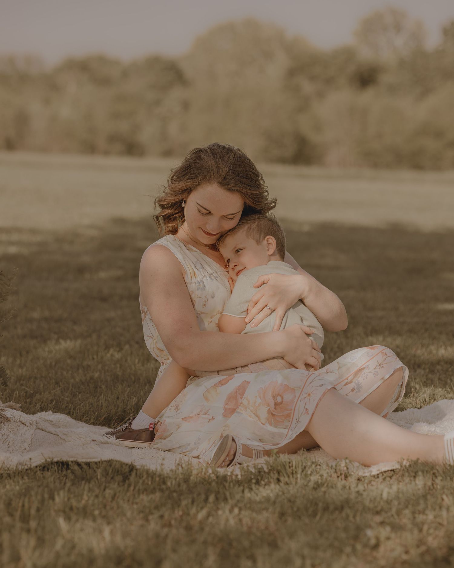 Mother tenderly cradling infant while sitting on grass in soft floral dress during warm outdoor photo session.