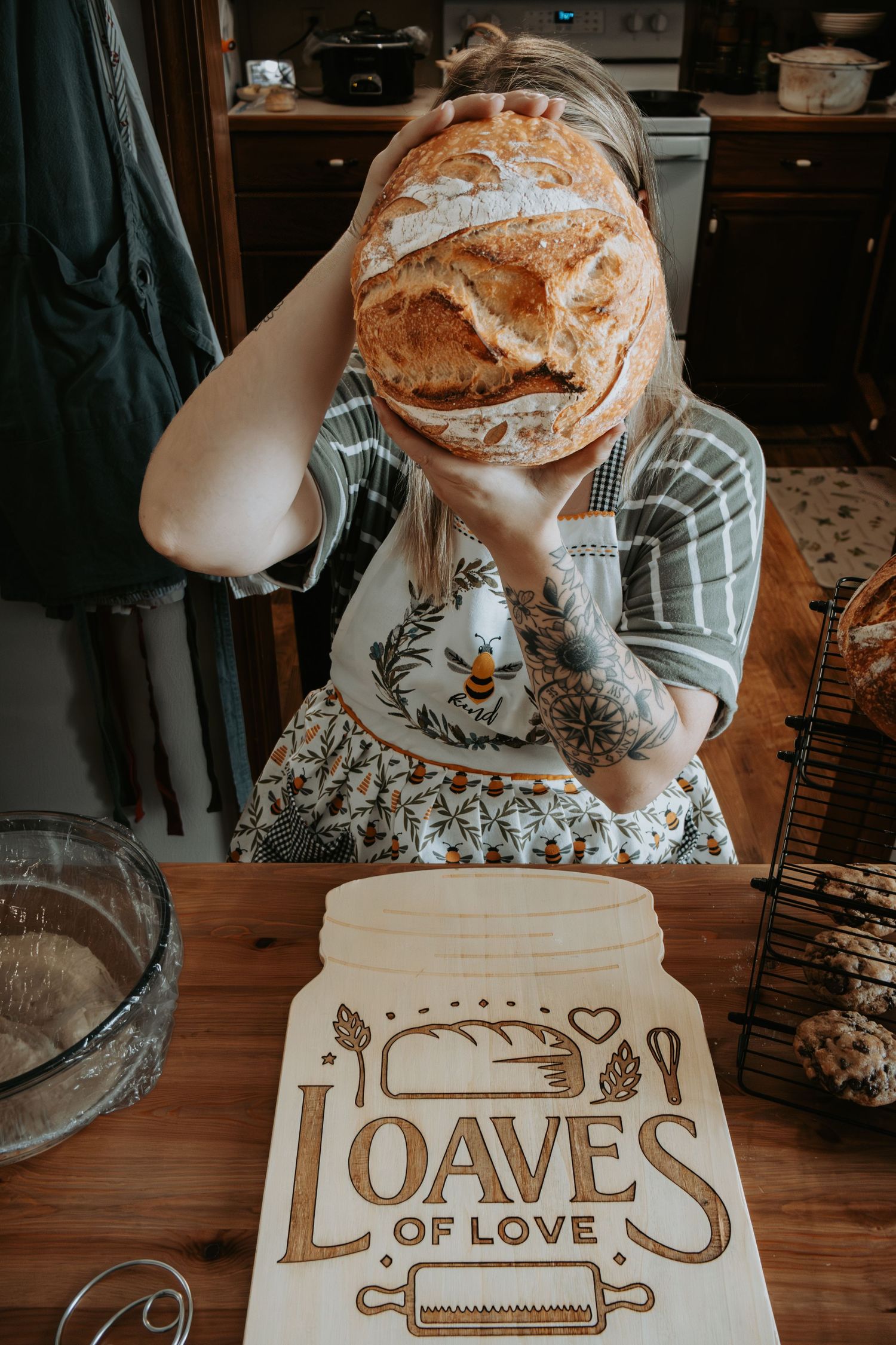 Woman holding a rustic sourdough loaf over her face at a wooden table with a Loaves of Love cutting board.