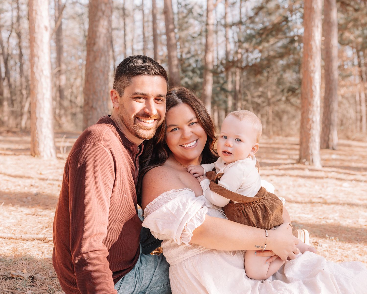 Happy young family with parents smiling and holding their baby outdoors in a sunny pine forest setting.