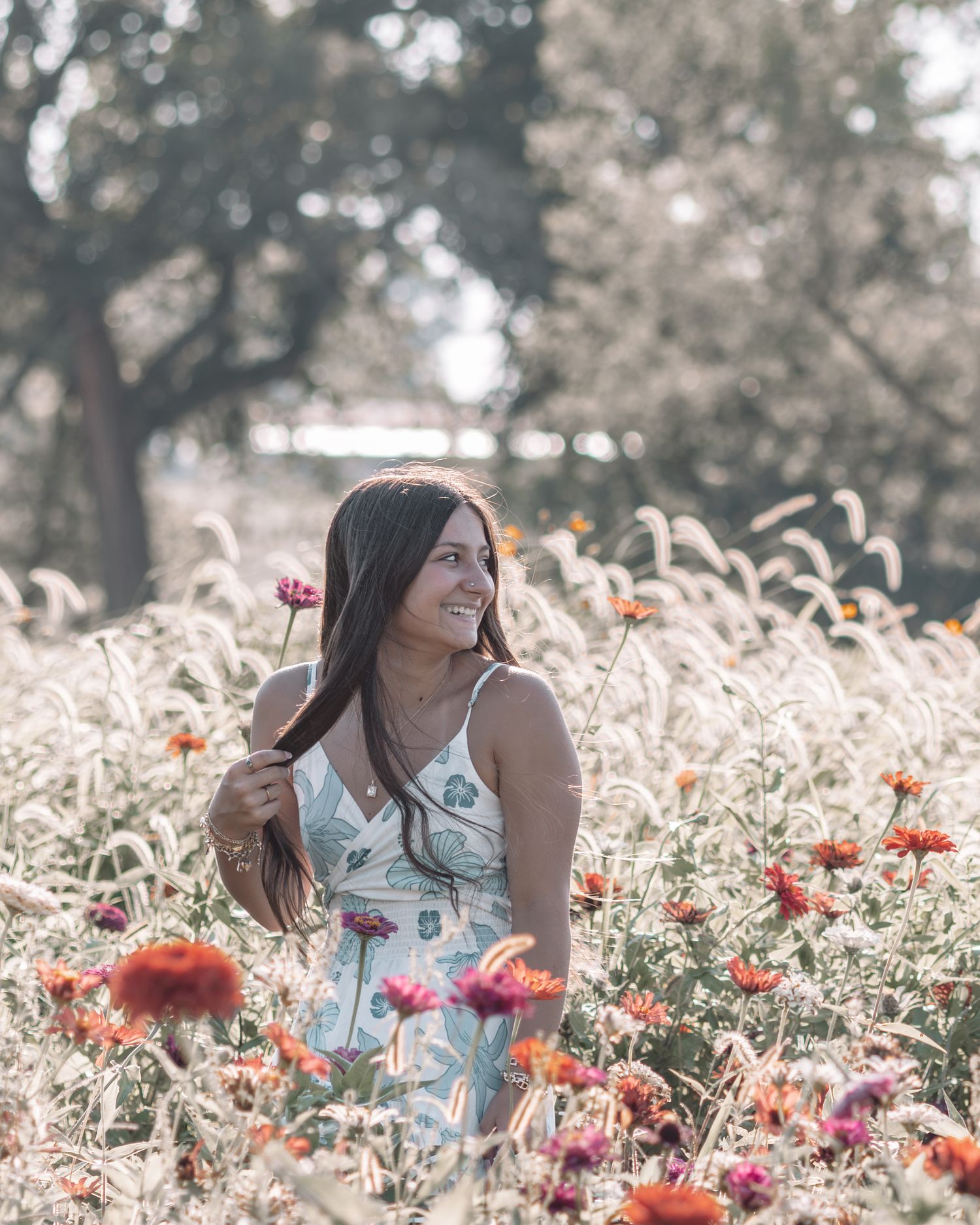 A smiling young woman in a floral dress stands among vibrant wildflowers in a sunlit meadow.