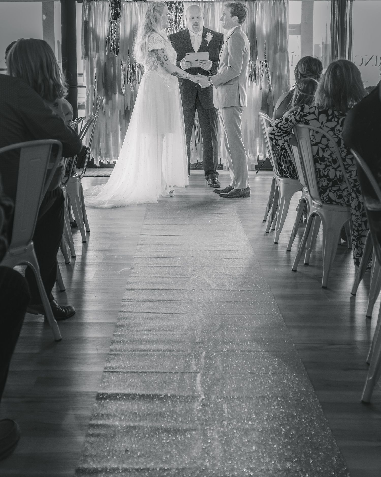 Black and white photo of bride and groom exchanging vows at indoor wedding ceremony with seated guests.