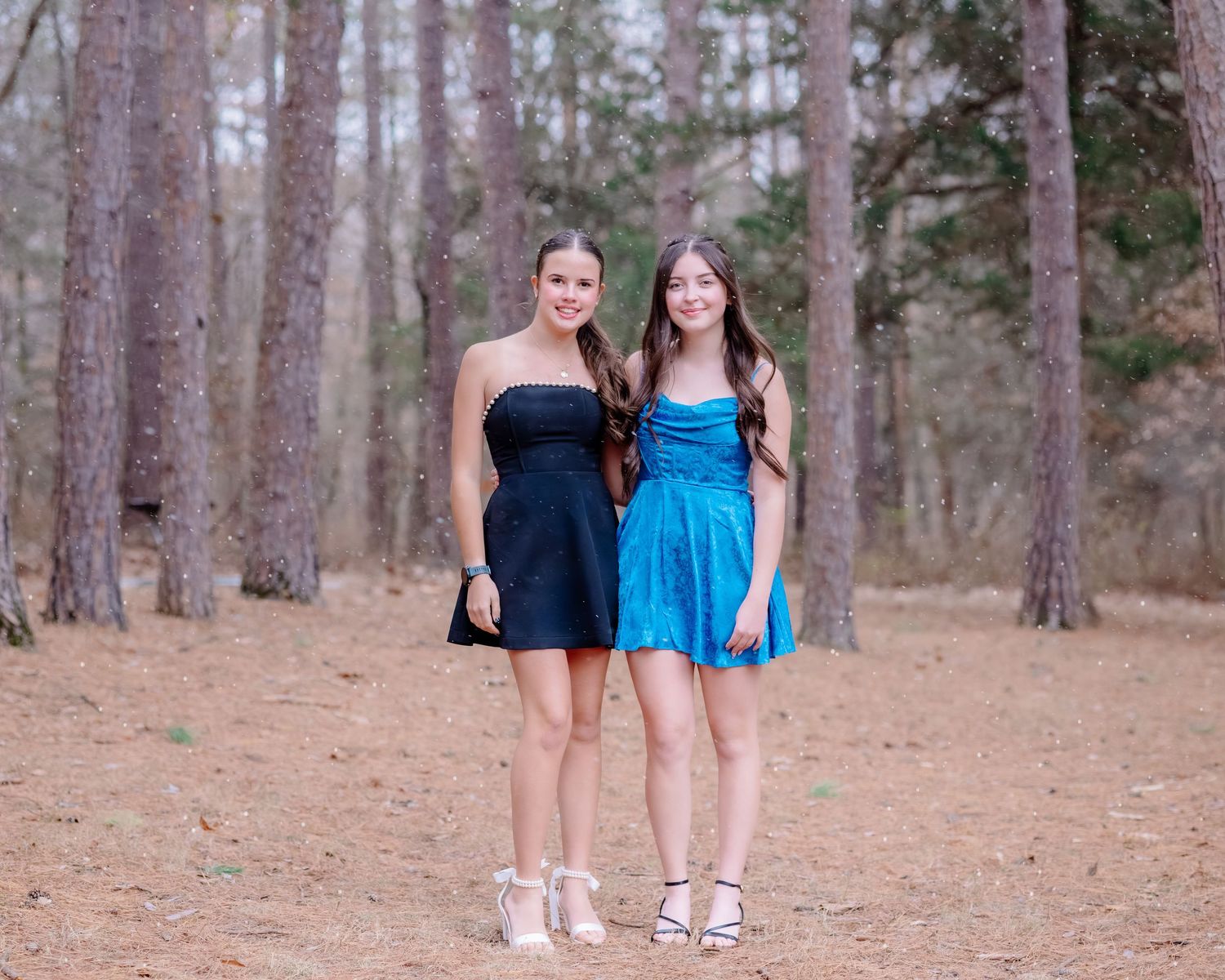 Two people in casual outfits stand together in a forest setting with pine trees and brown ground cover.