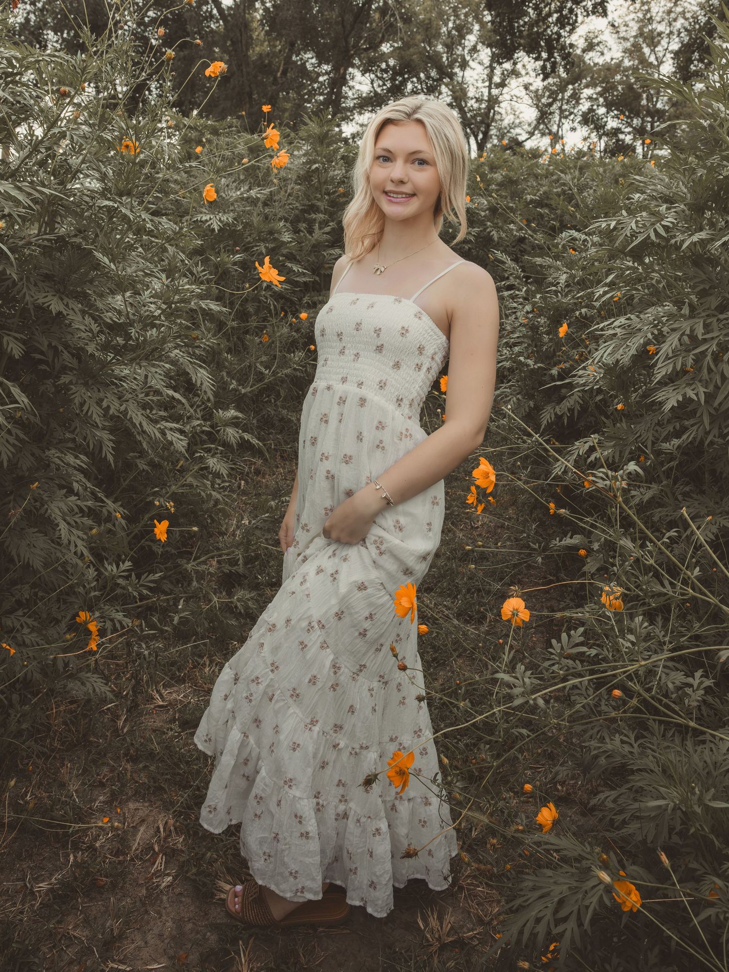 Blonde woman in floral maxi dress standing among orange wildflowers and lush greenery outdoors.