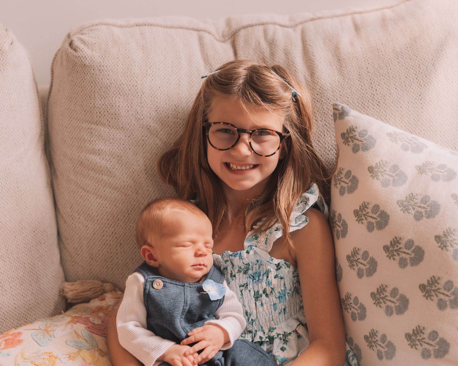 Young girl with glasses holding a sleeping newborn baby on a cozy beige couch with floral pillows.