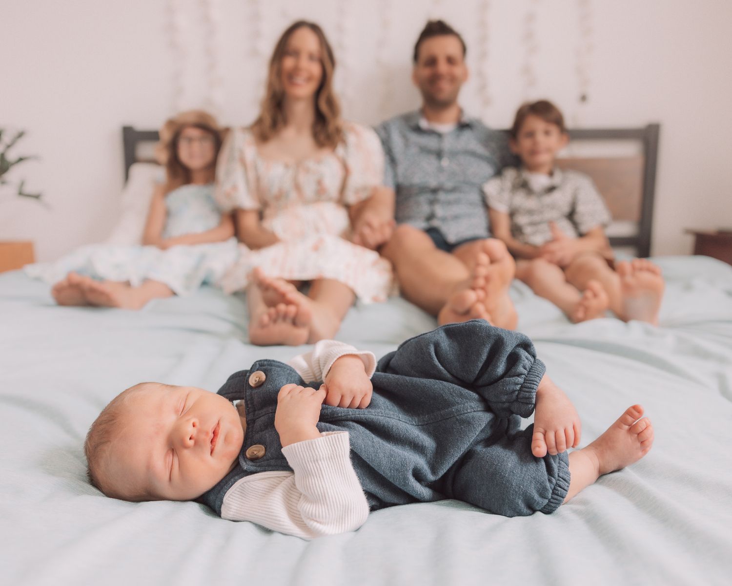 Family of five sitting on a white bed with newborn baby boy in blue overalls in the foreground.