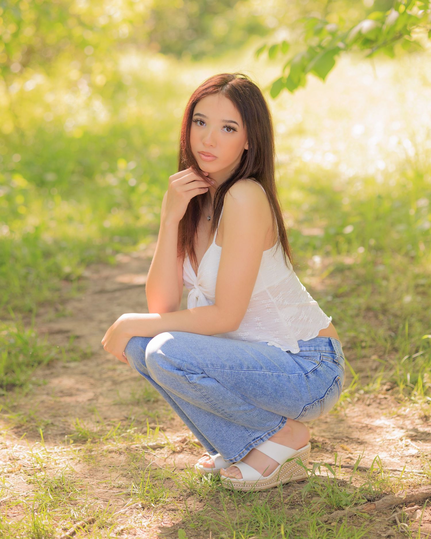 Young woman in white top and jeans crouching on a sunlit forest path, posing for a senior portrait photo.