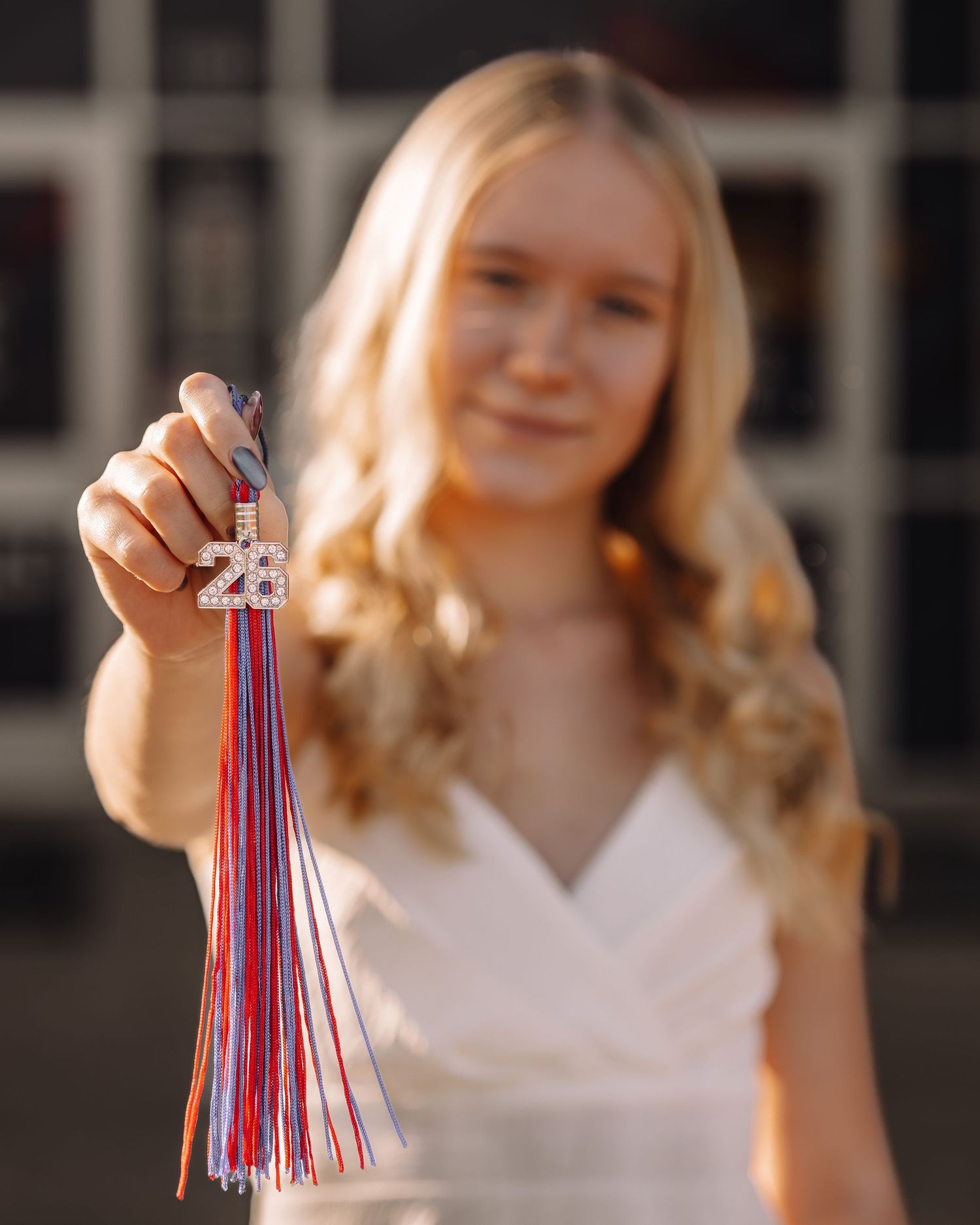 Blonde girl holding a rhinestone 2023 graduation tassel charm with red, white, and blue ribbons for senior photos.