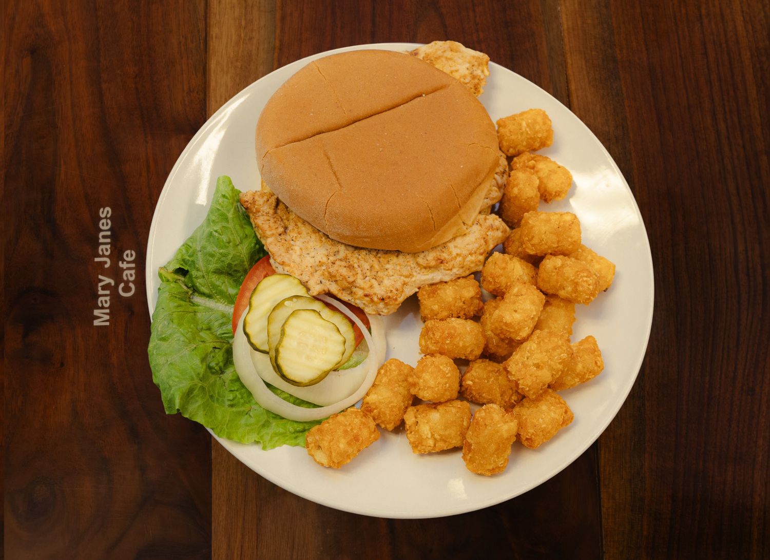 Crispy chicken sandwich with lettuce, onion, and pickles on a bun, served with golden tater tots on a white plate.