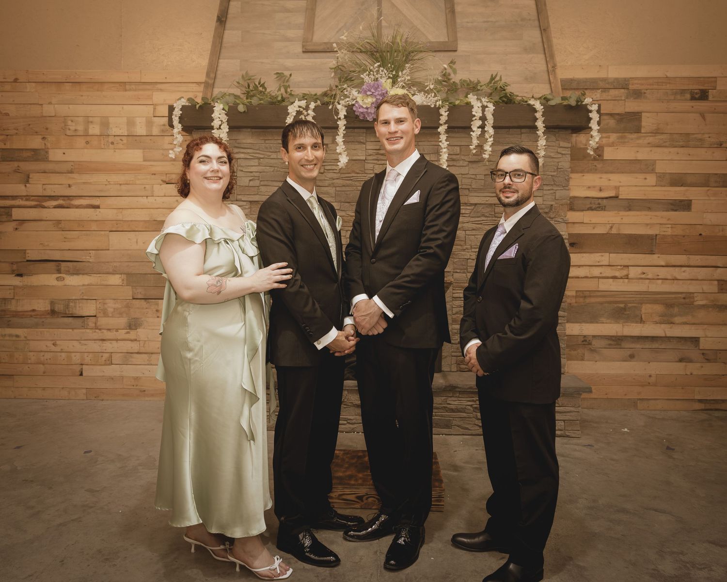 Four wedding guests pose in front of rustic wood plank wall with floral garland at indoor venue.