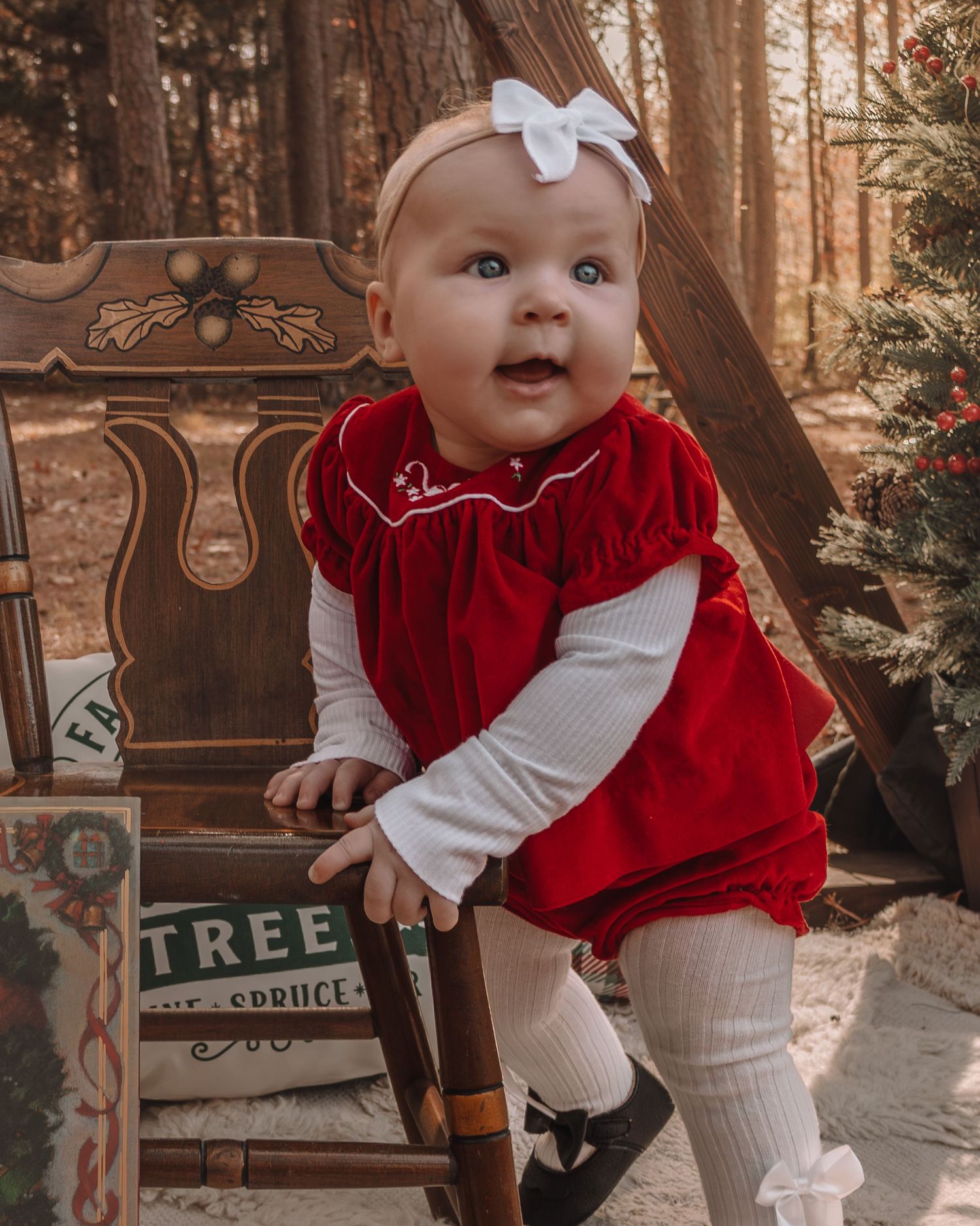 A small child in a festive red velvet dress and white bow stands by a vintage Christmas tree sign in a forest setting.