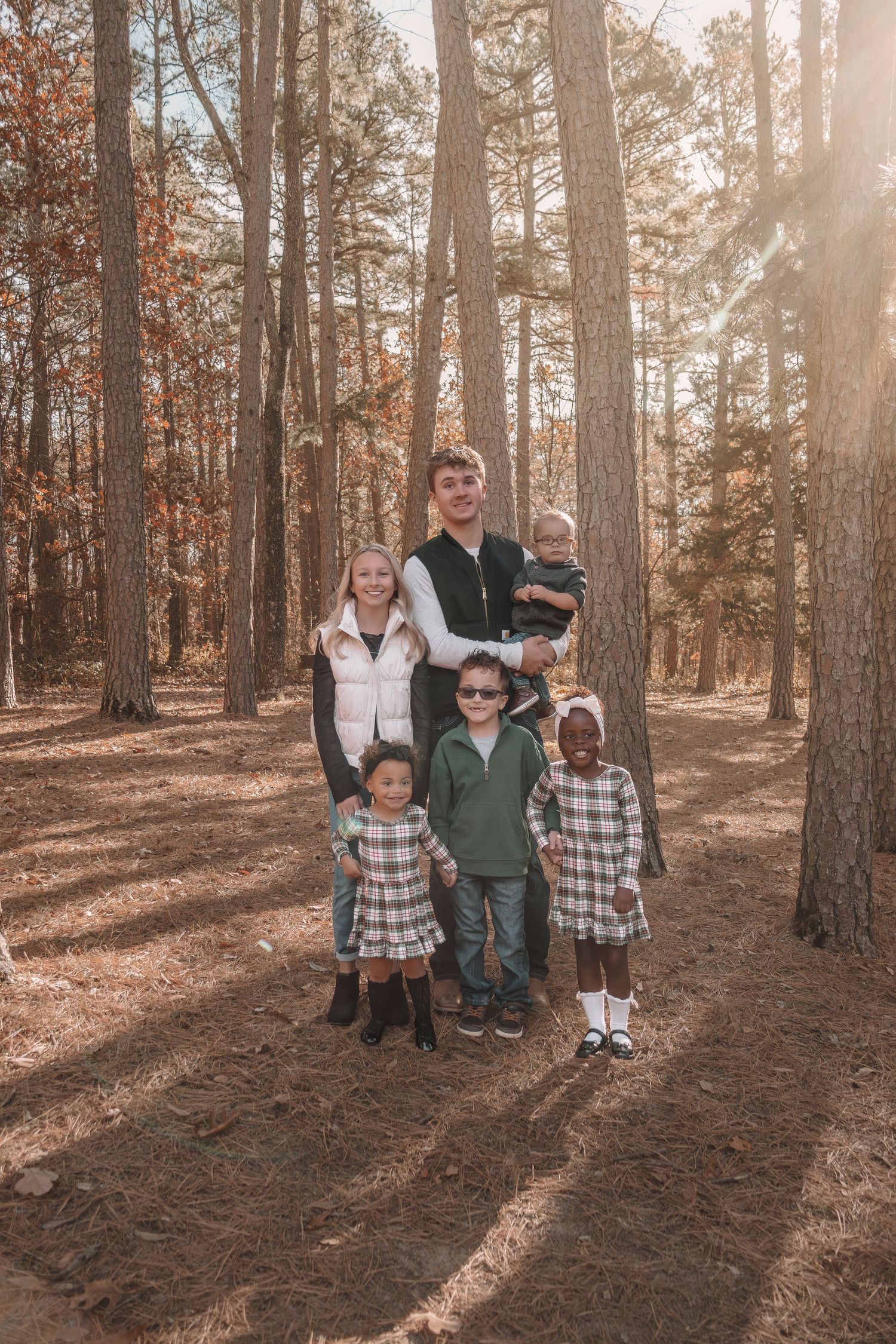A family poses together in a sunlit autumn forest with pine trees and fallen leaves creating a warm atmosphere.