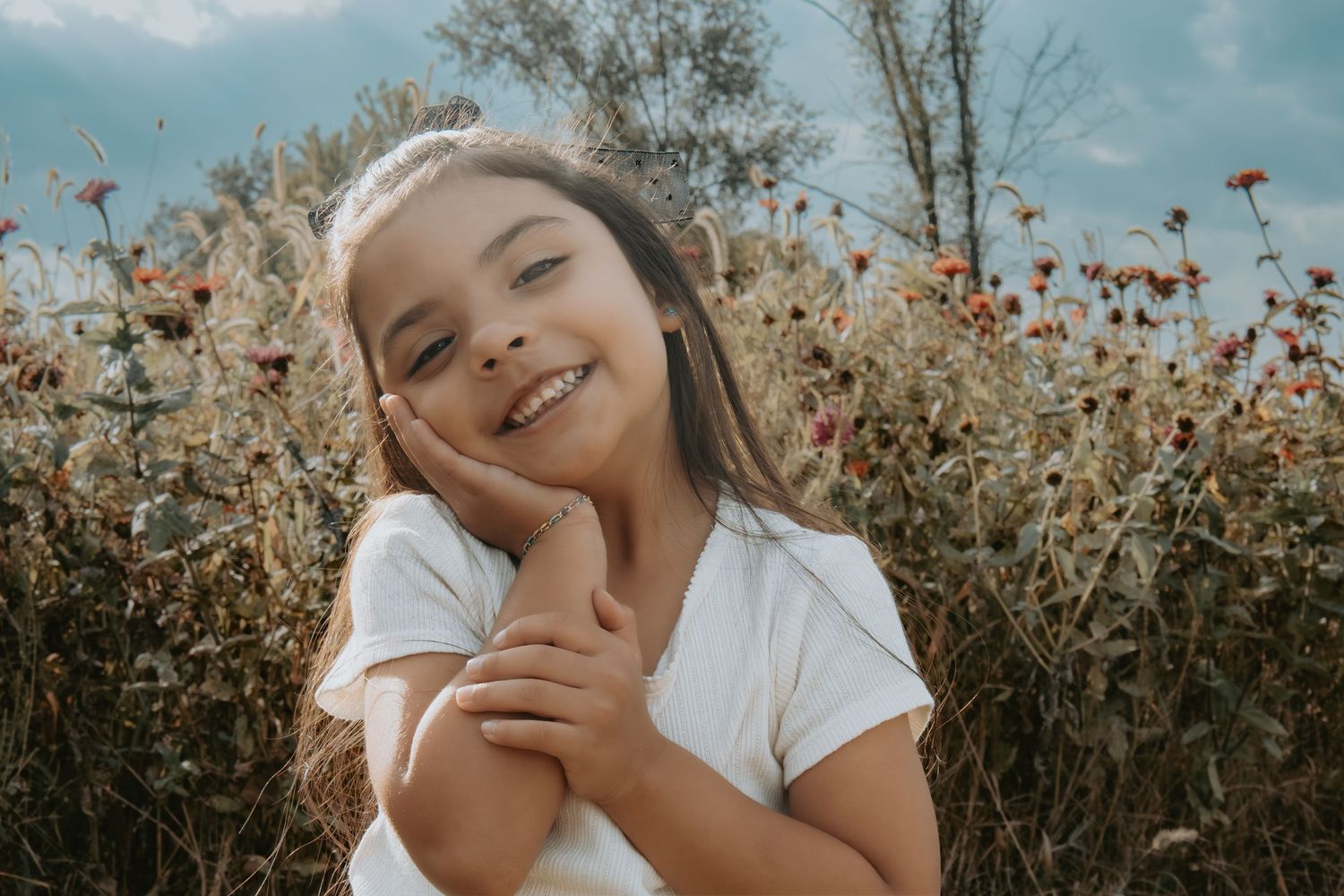 A smiling young girl with long hair poses cheerfully in a white t-shirt among wildflowers in a sunny field.