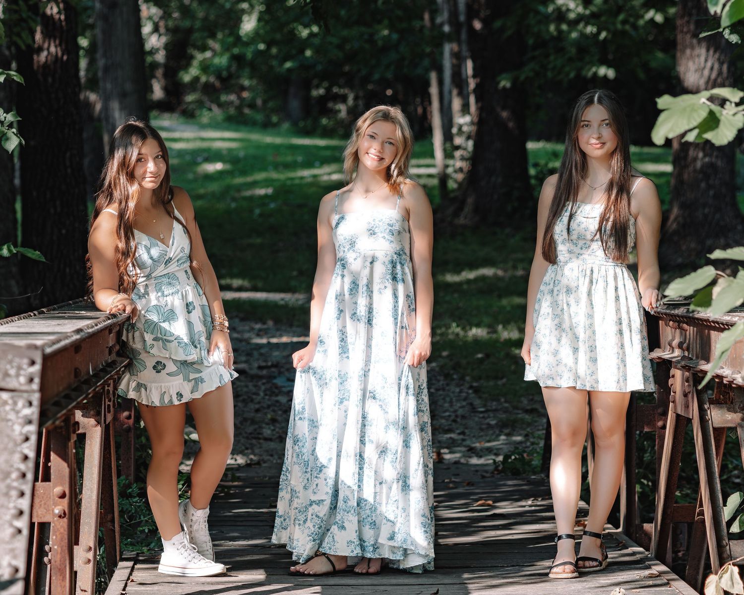 Three friends wearing light floral summer dresses pose together on a wooden deck in a sunlit wooded setting.