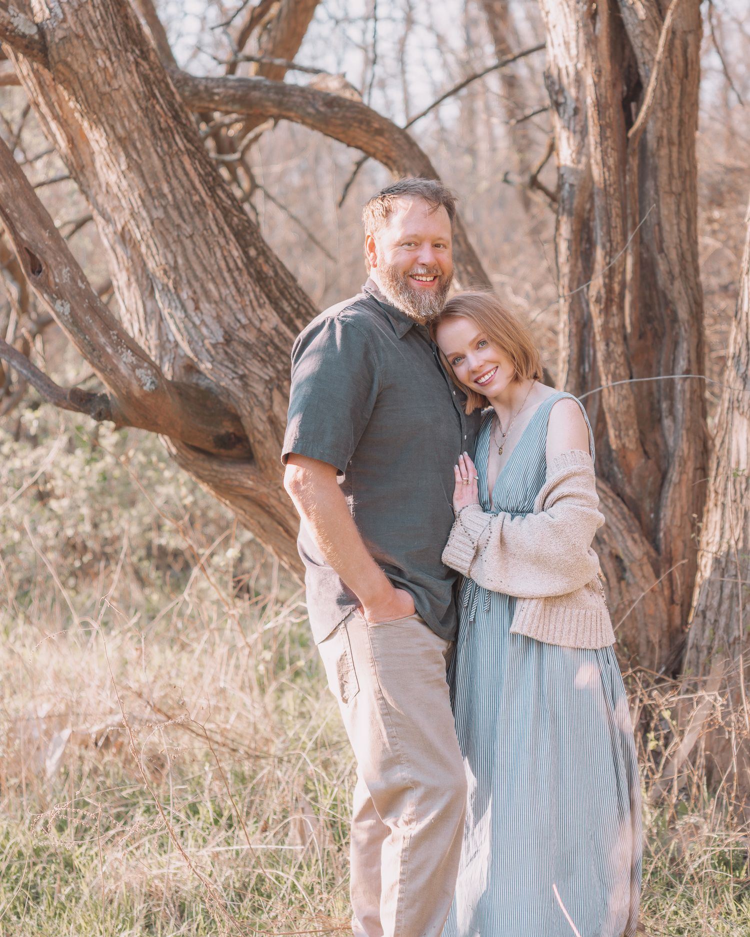 A couple poses warmly in a sunlit field with bare trees, the woman in a teal dress leaning on the mans shoulder.