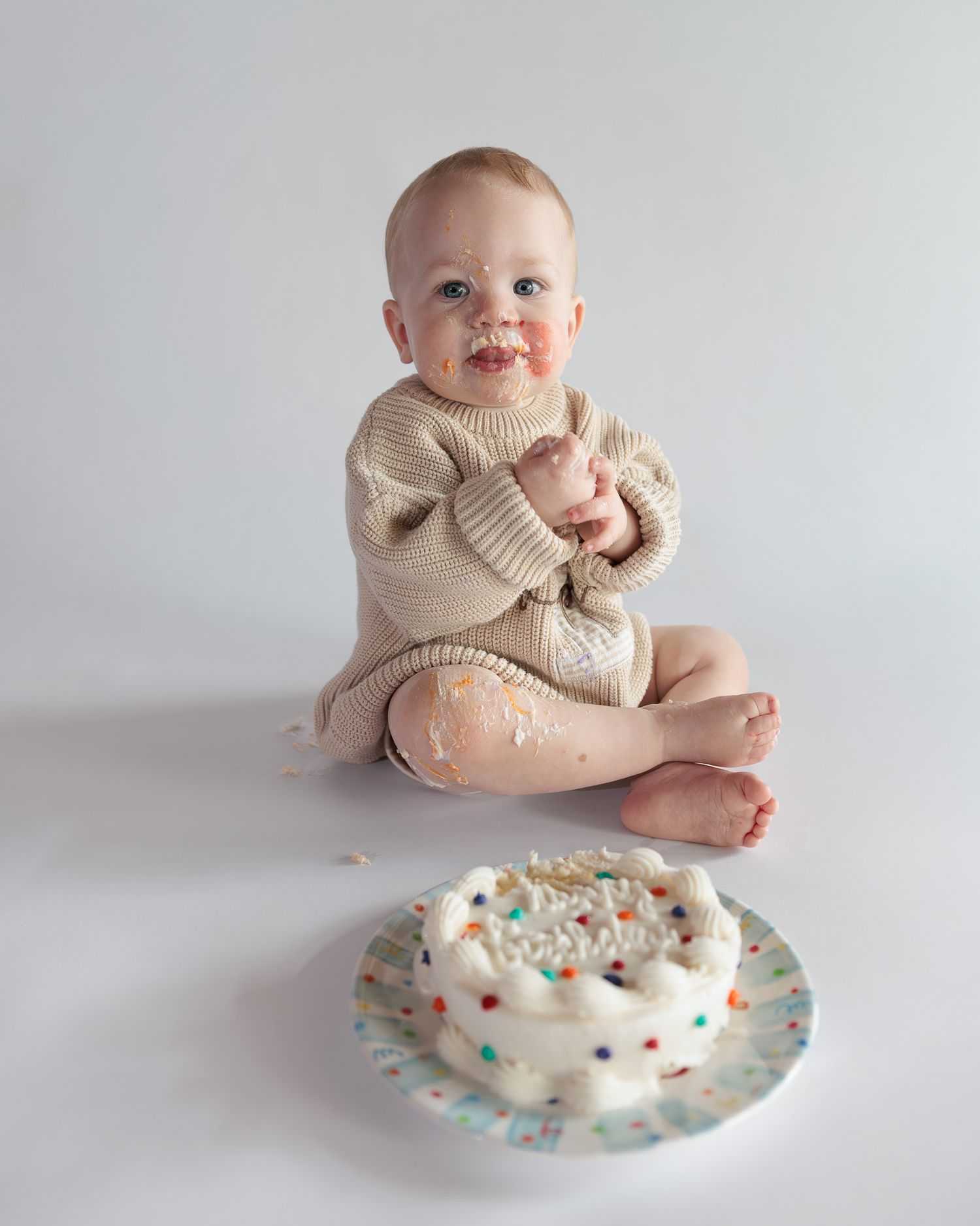 Baby with frosting on face sits next to a smash cake with colorful sprinkles during first birthday celebration.