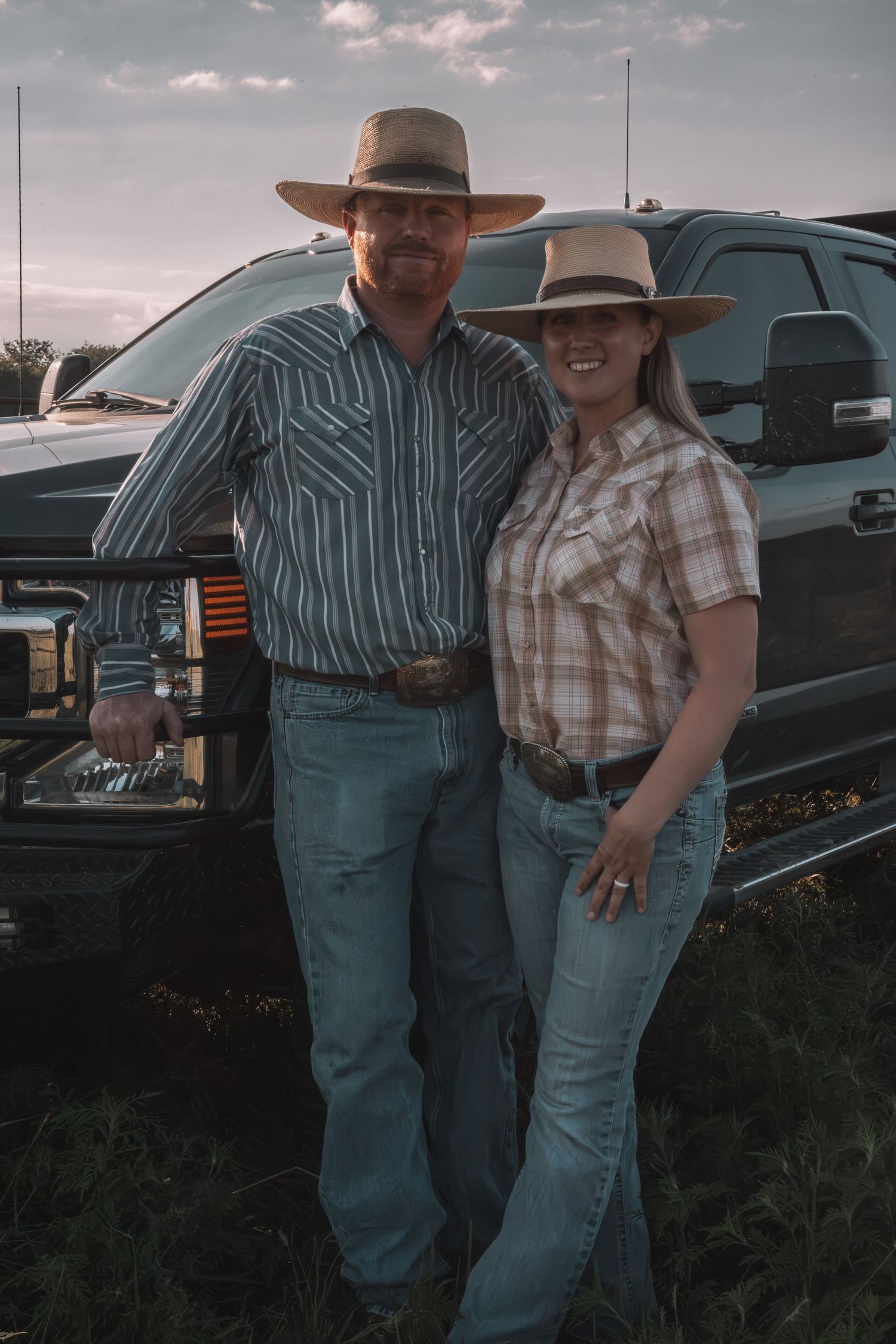 A couple in cowboy hats and western shirts pose smiling in front of a black pickup truck at dusk.
