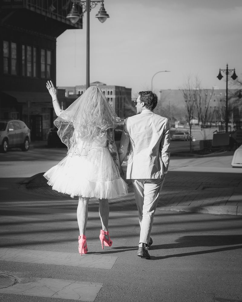 A couple walking down a street, with the person in white dress wearing bright pink heels in black and white photography.
