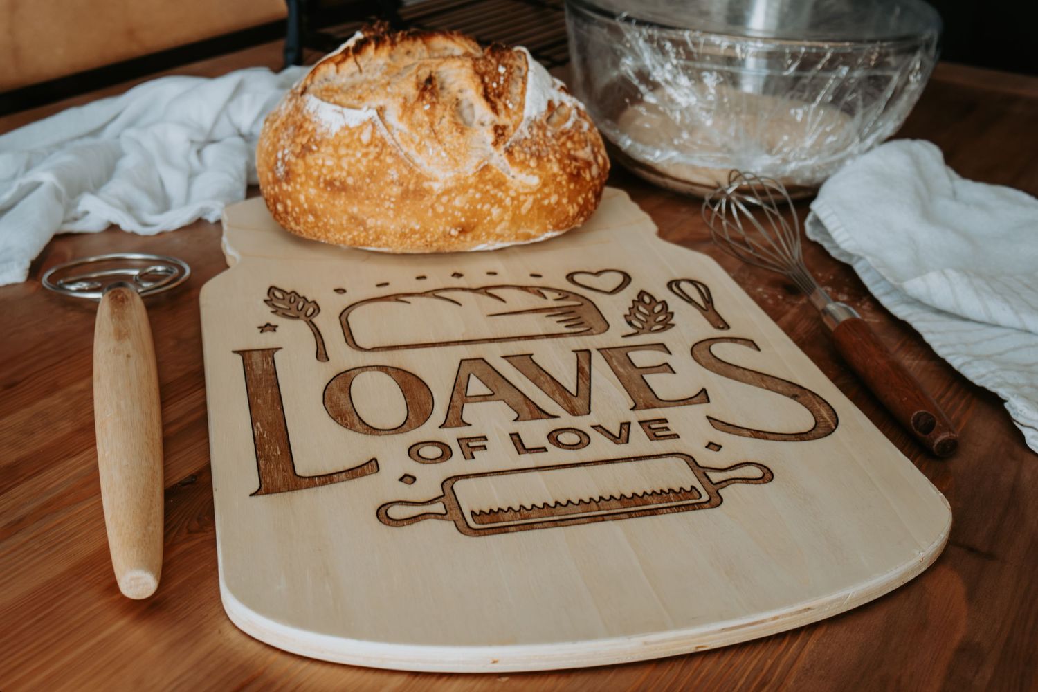 Wooden cutting board engraved with Loaves of Love text, displayed with artisan bread, rolling pin, and whisk.